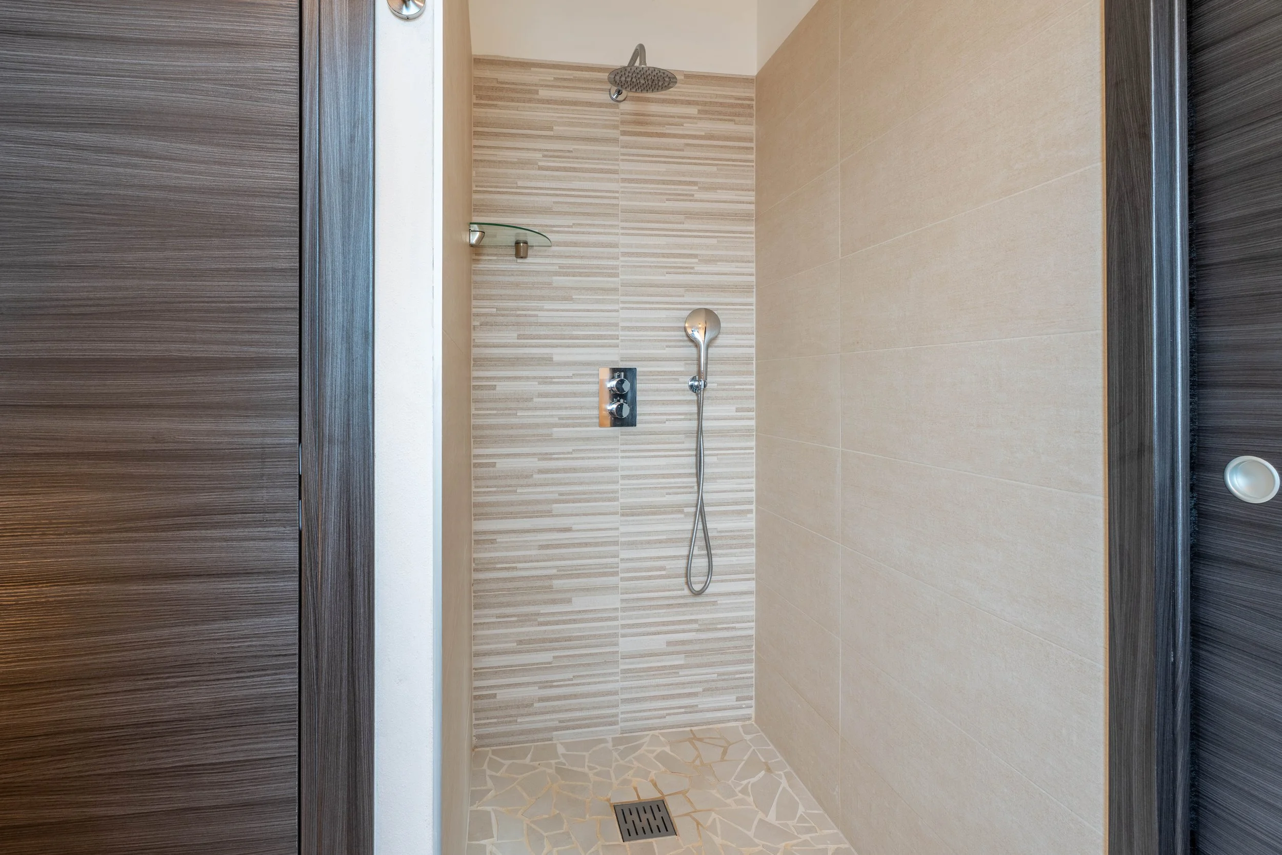 Inrichting van een moderne douche met beige en stenen tegels, een hoofd douche en een handdouche, kleine glazen plank in de hoek.