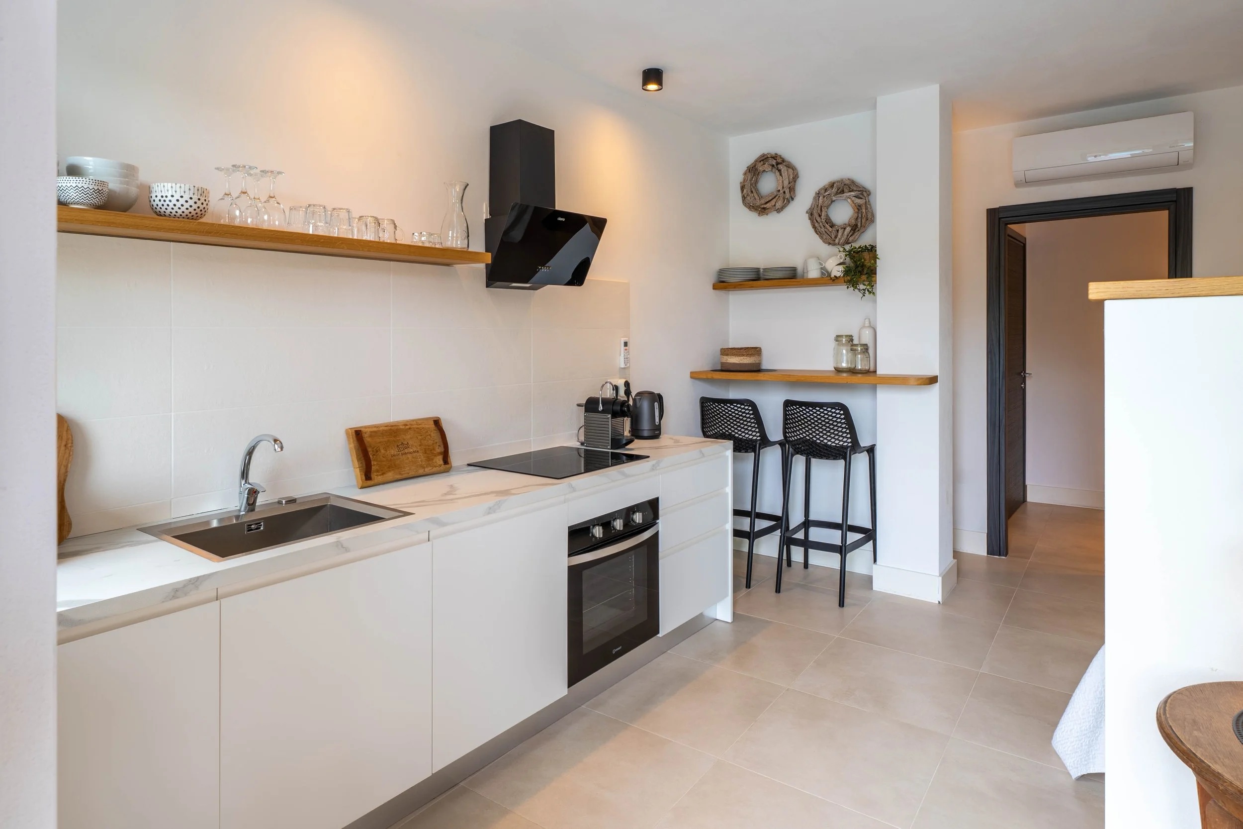 Kleine moderne keuken met witte kasten, een oven, een inductiekookplaat, open houten planken met keukengerei en decoratie, een hardhouten bartafel met twee zwarte krukken, en een airconditioning unit.