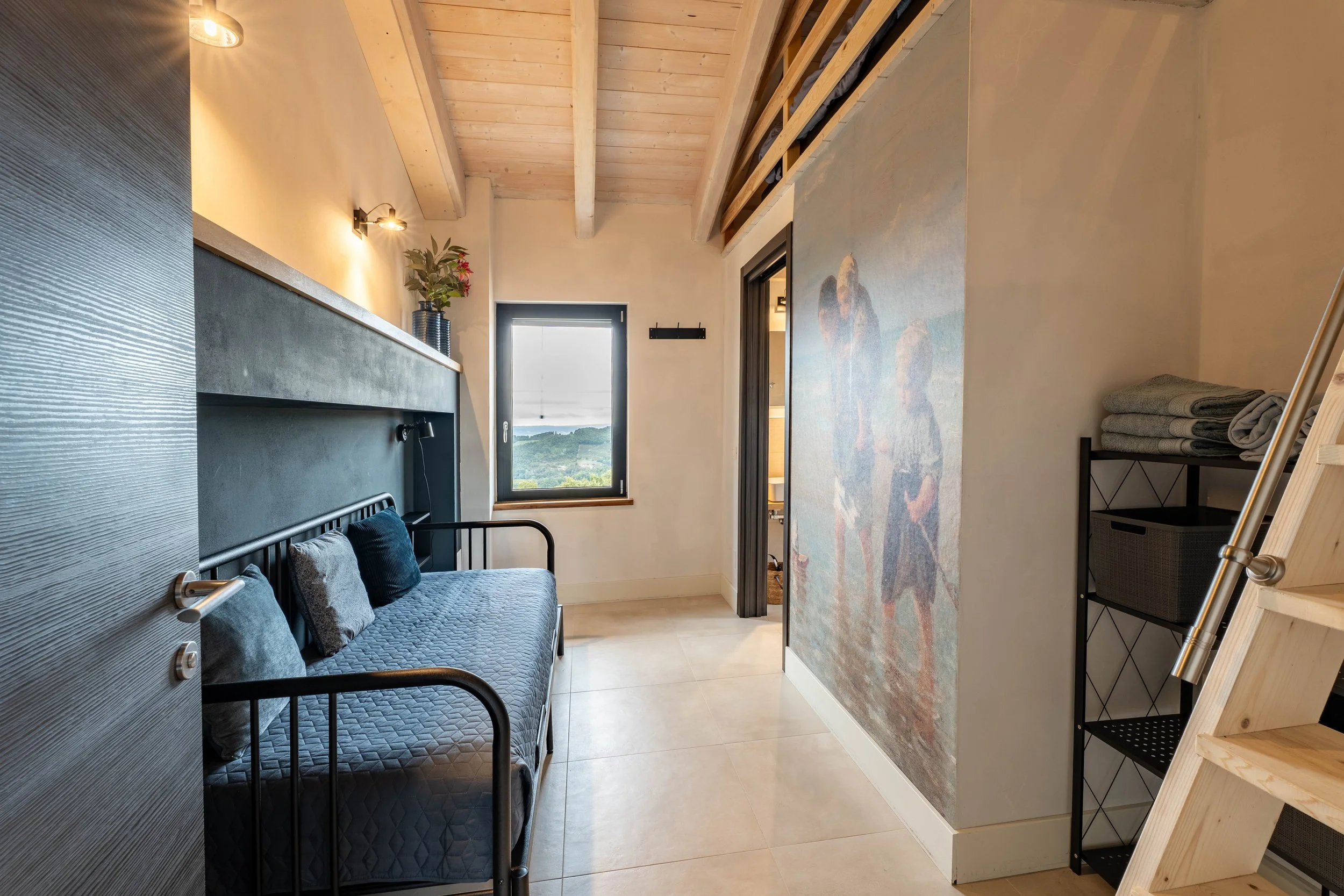 Kleine slaapkamer met houten plafond, zwart metalen bed met grijze beddengoed en kussens, muur met muurschildering van een vrouw en een kind, venster met uitzicht op groene heuvels, open deur naar badkamer, metalen plank met handdoeken en een opklapbare ladder.