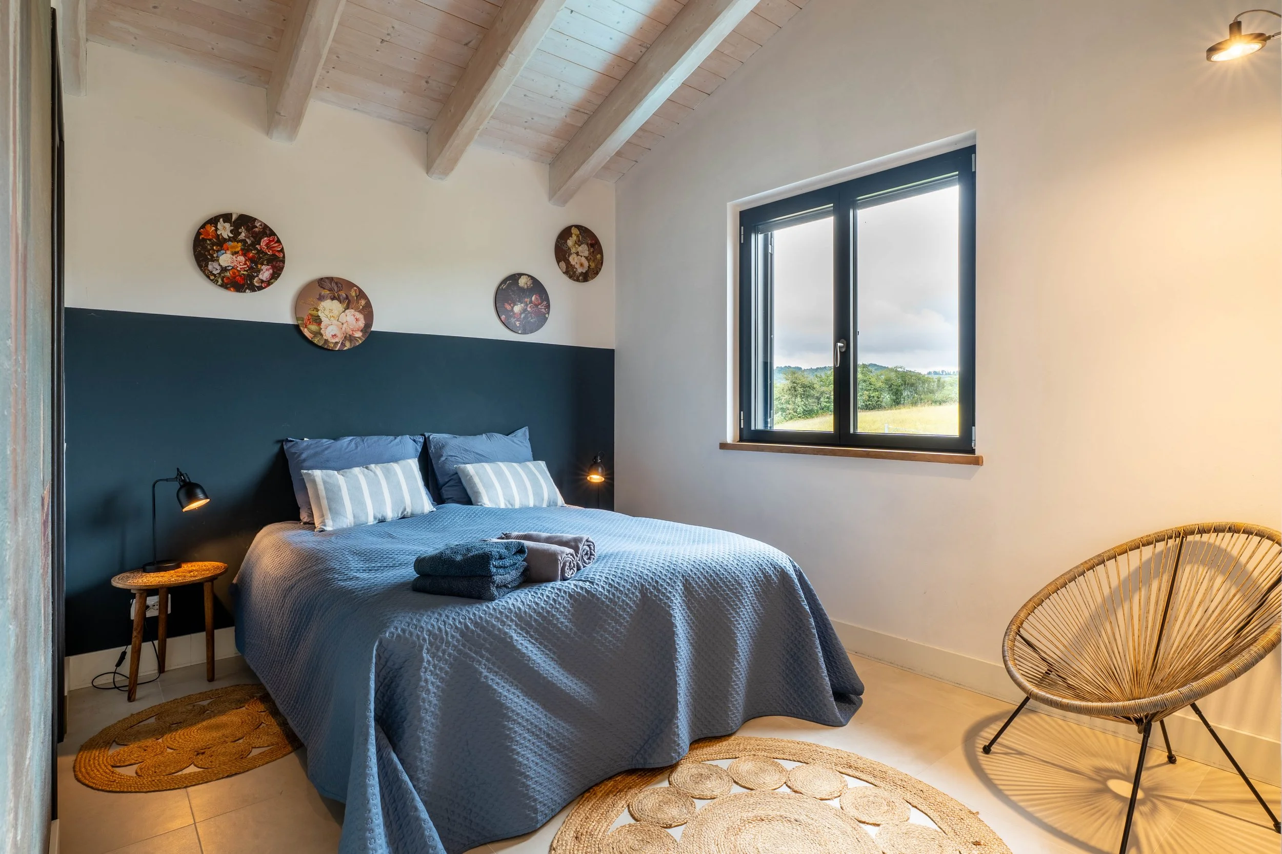 Slaapkamer met blauwe beddengoed, houten plafond met witte balken, muur met bloemen decoratie, raam met uitzicht op groen landschap, rond houten tapijt en een rieten stoel.