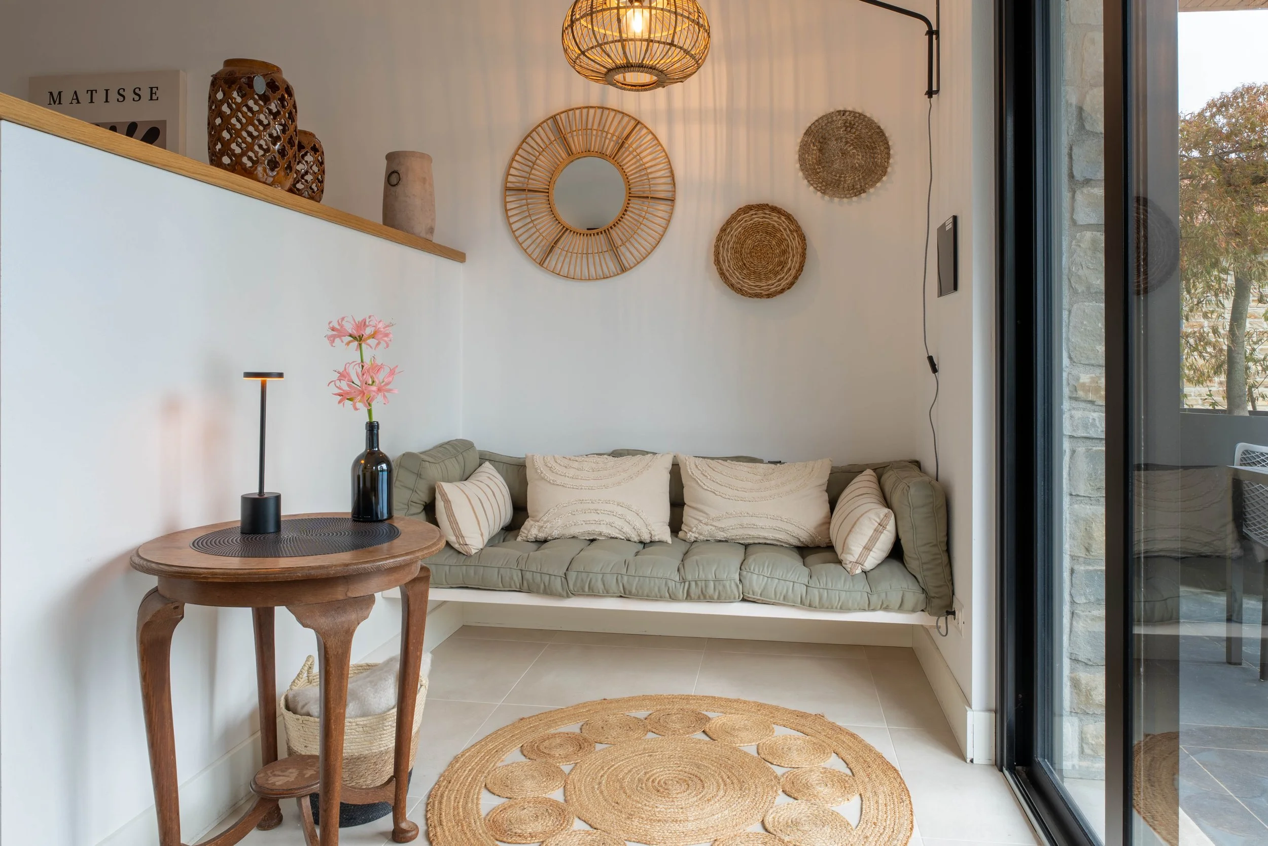 Een gestoffeerde bank met kussens, een ronde houten salontafel met een zwarte lamp en vaas met roze bloemen, een rotan vloerkleed, en muurdecoratie van manden en een spiegel in een lichte woonkamer met grote raam en natuurlijke decoratie.