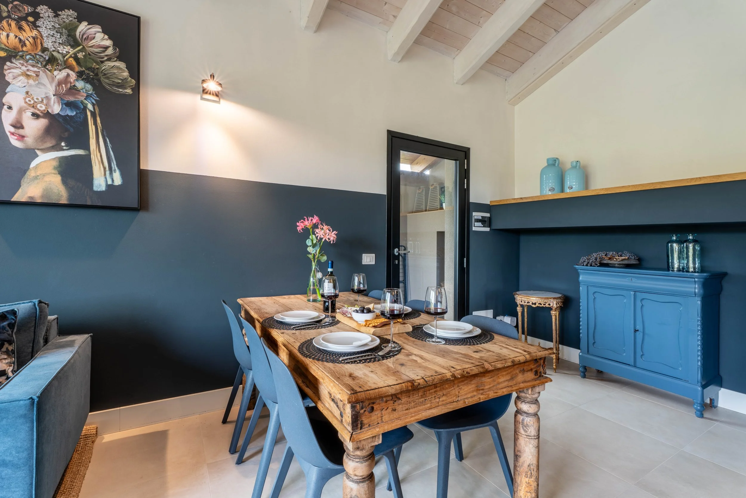 Een eethoek met een rustieke houten tafel, zes blauwe stoelen, een vaas met bloemen, wijn- en delicatessen op de tafel, een blauwe kast, en muurdecoratie inclusief een schilderij van een meisje in een kleurrijk kostuum, binnen in een woning met een schuine houten plafond.