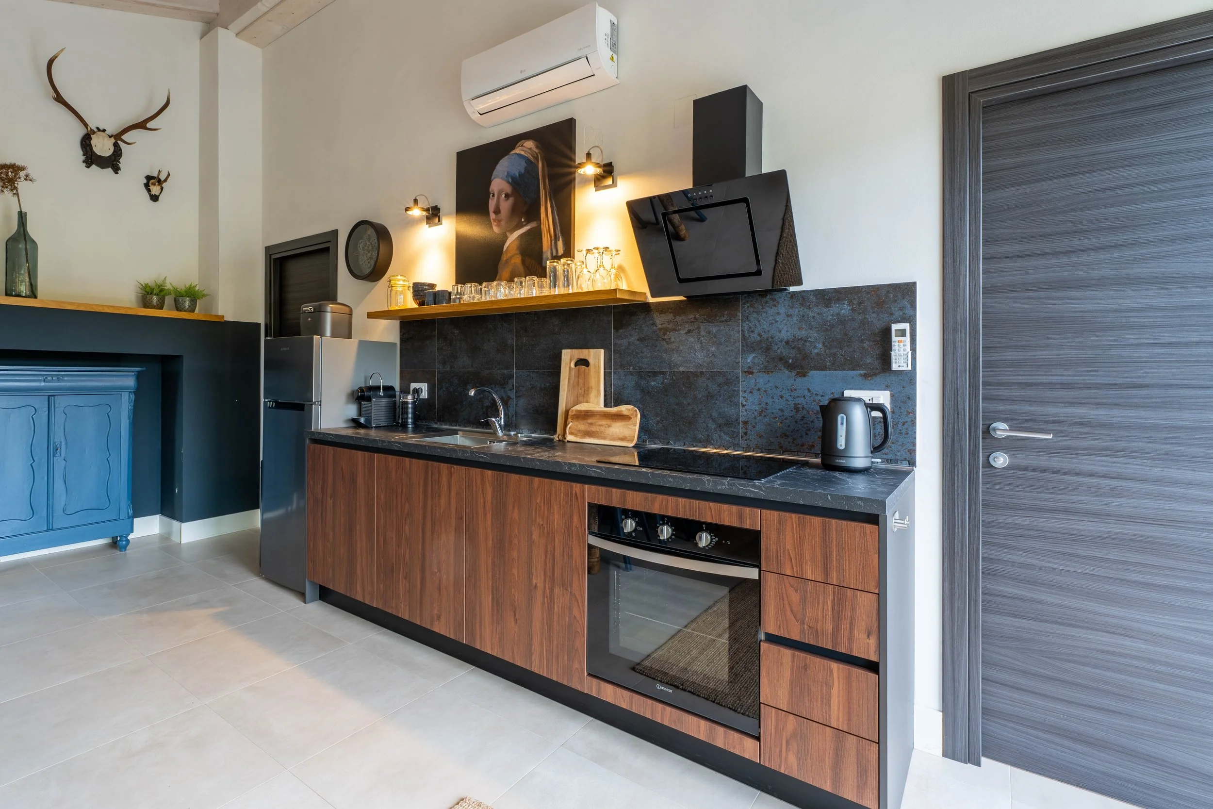 Een moderne keuken met houten kasten, zwart blad en donker stenen achterwand, met verschillende kleine keukenapparaten, een koelkast en decoratie.