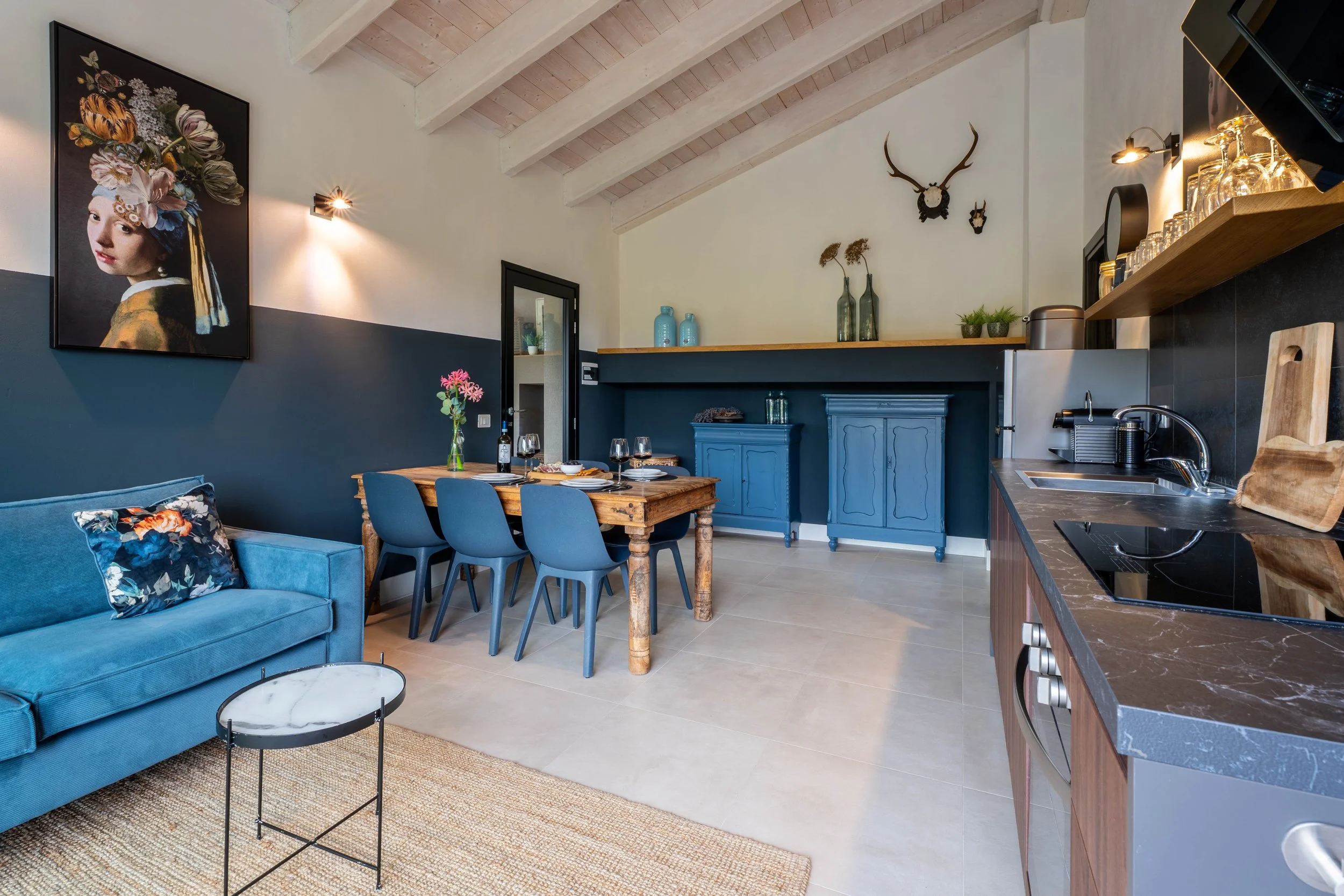 Een moderne open keuken en eethoek met een blauwe bank, een kunstwerk van een vrouw met bloemen in haar haar, en decoratieve elementen zoals een geweer en planten. Zeer sfeervolle en stijlvolle inrichting.