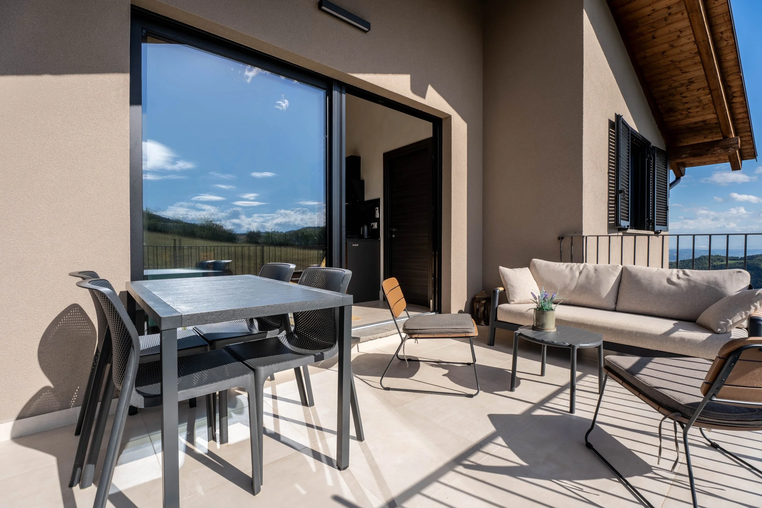 Buitenpatio met tafel, stoelen, bank en plant, uitzicht op landschap en blauwe lucht met wolken.