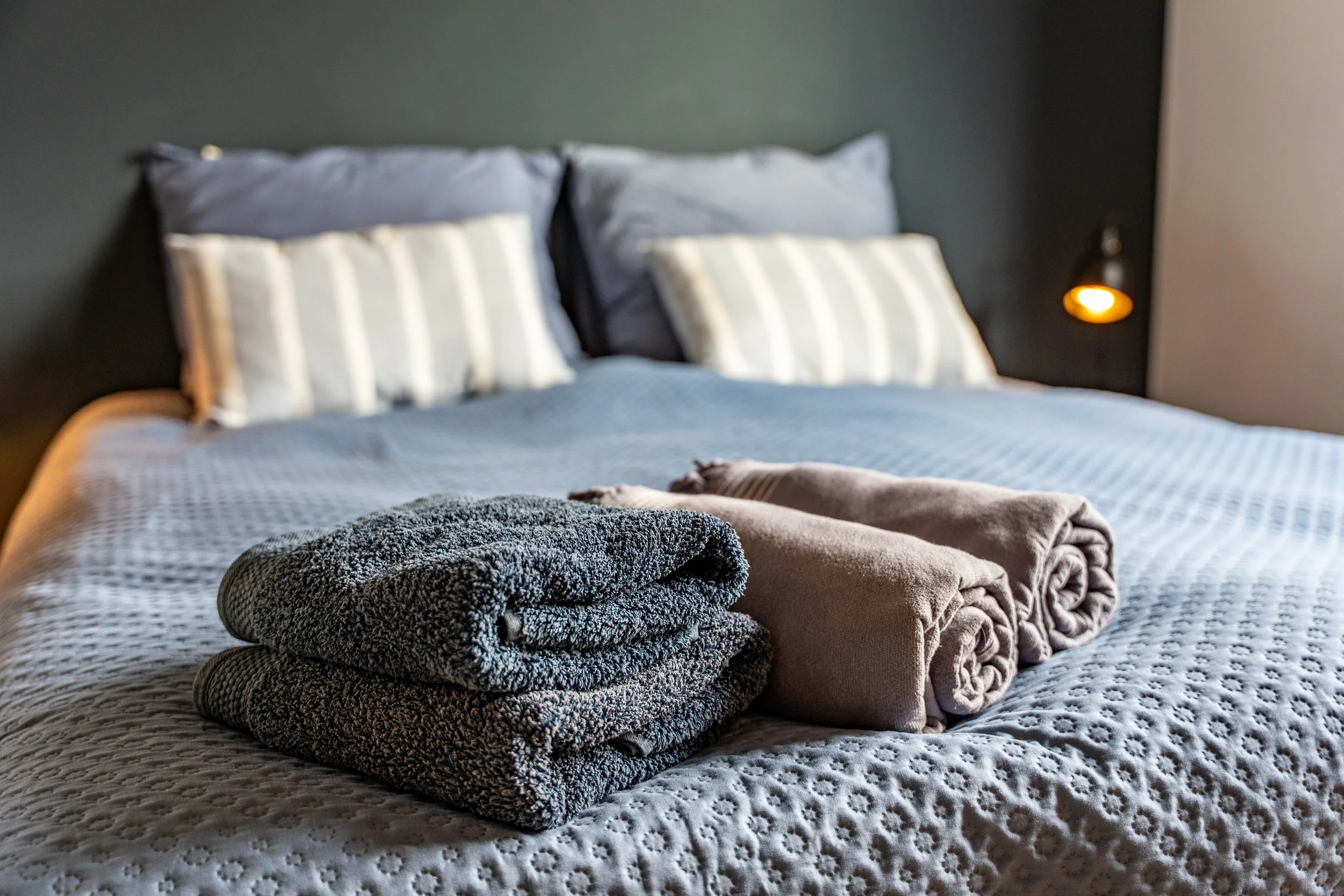 Op een bed liggen twee gedraaide handdoeken en twee badjassen. De achtergrond toont een hoofdeinde met vier kussens en een muur met een leeslamp.