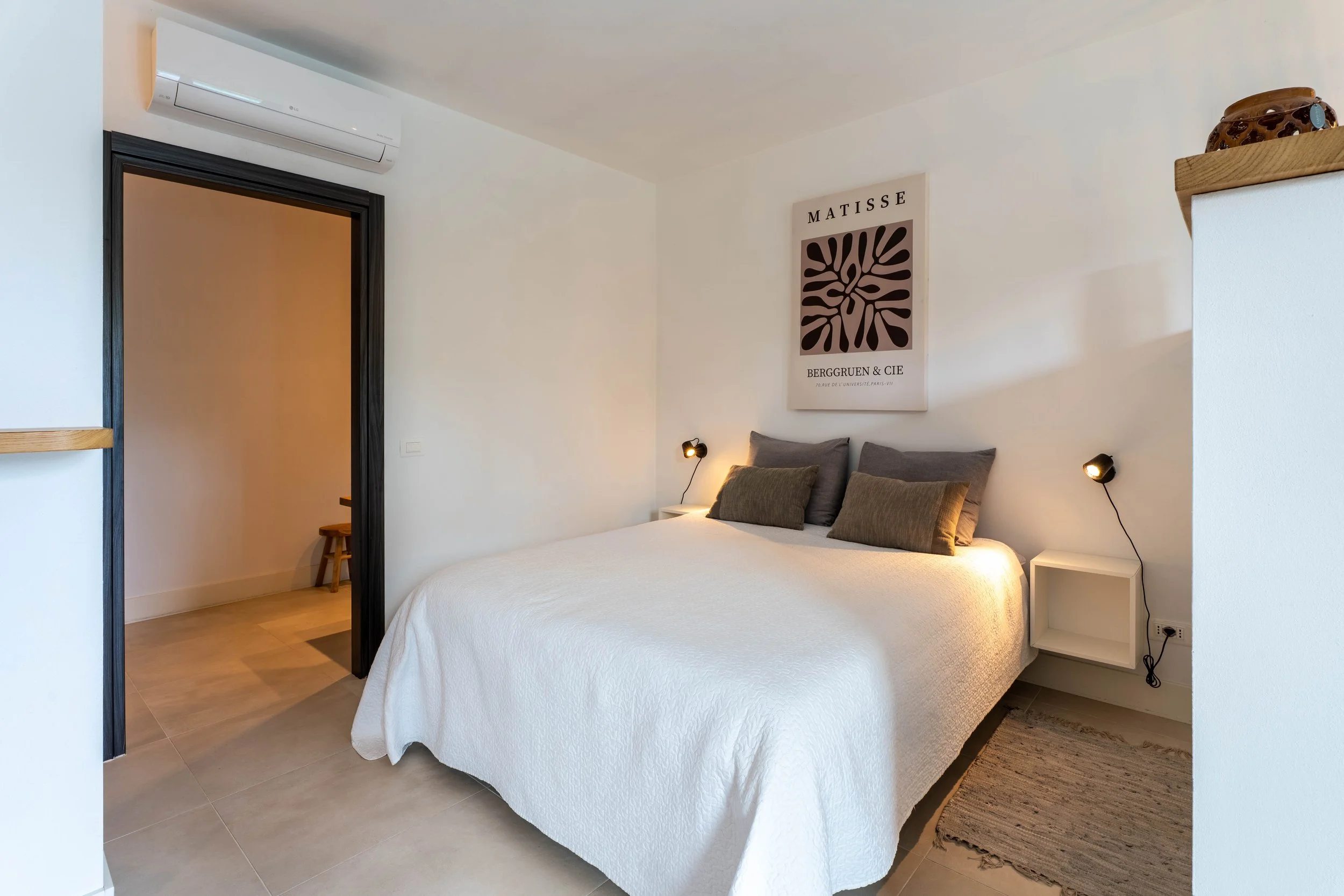 Slaapkamer met wit beddengoed, vier kussens, muurdecoratie van Matisse, muurplanten en een kleine tafel naast het bed, met moderne lampen aan de muur.