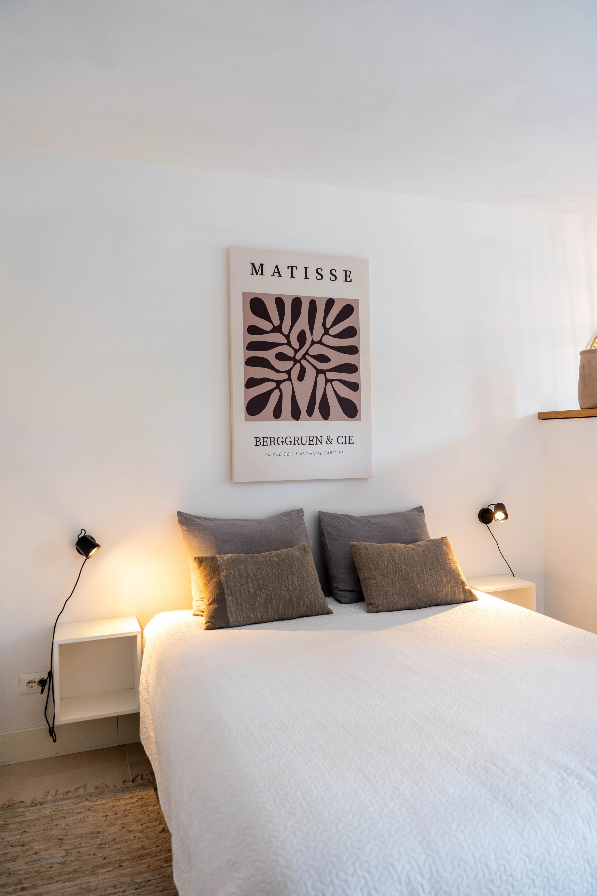 Een slaapkamer met een wit bed, vier kussens en twee leeslampjes aan de zijkanten, een poster van Matisse aan de muur en een ramenbank met manden.