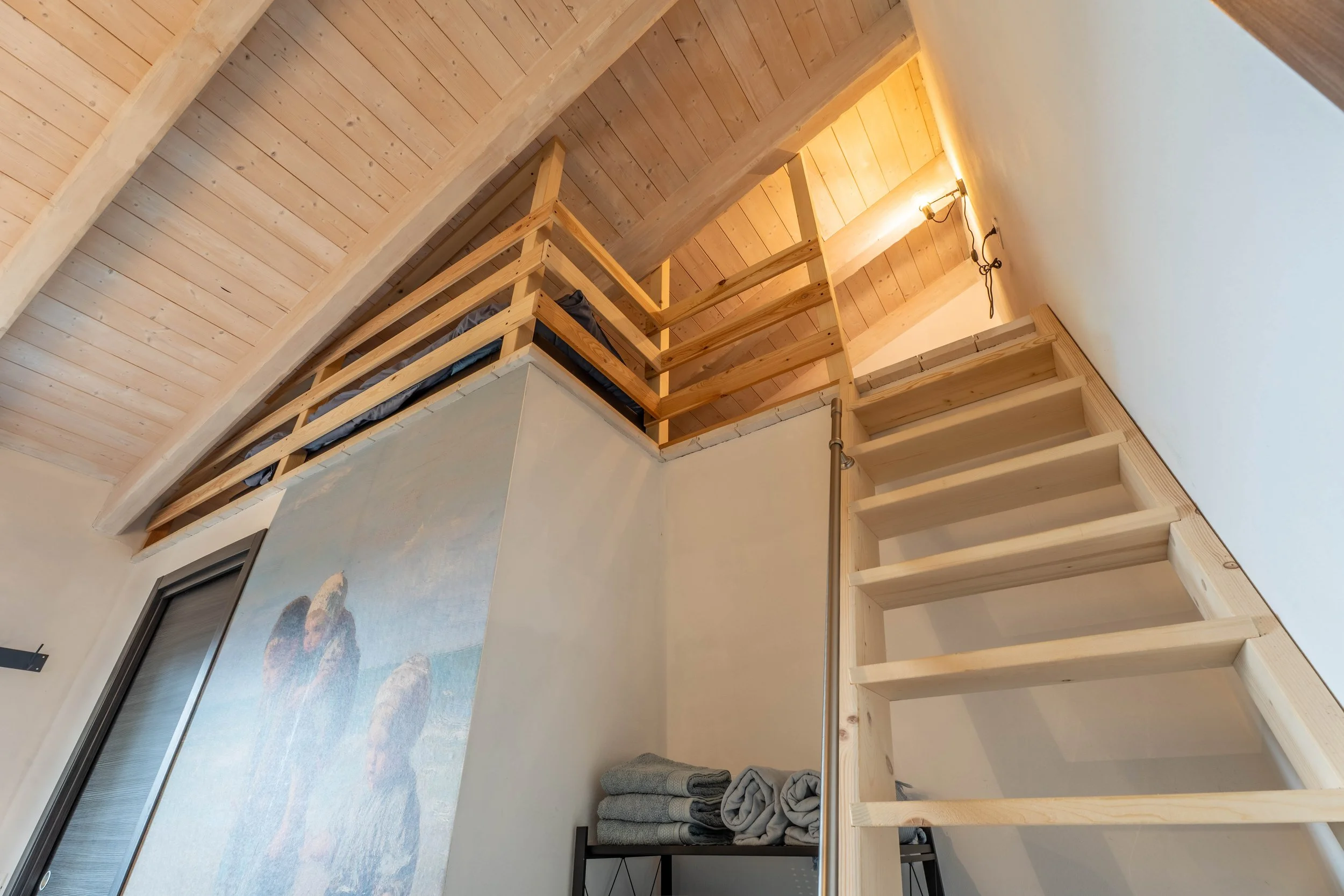Interieur van een woning met een leuning en trap in hout, een muur met een muurschildering van bergen en een raam, en een stapel handdoeken op een plank.