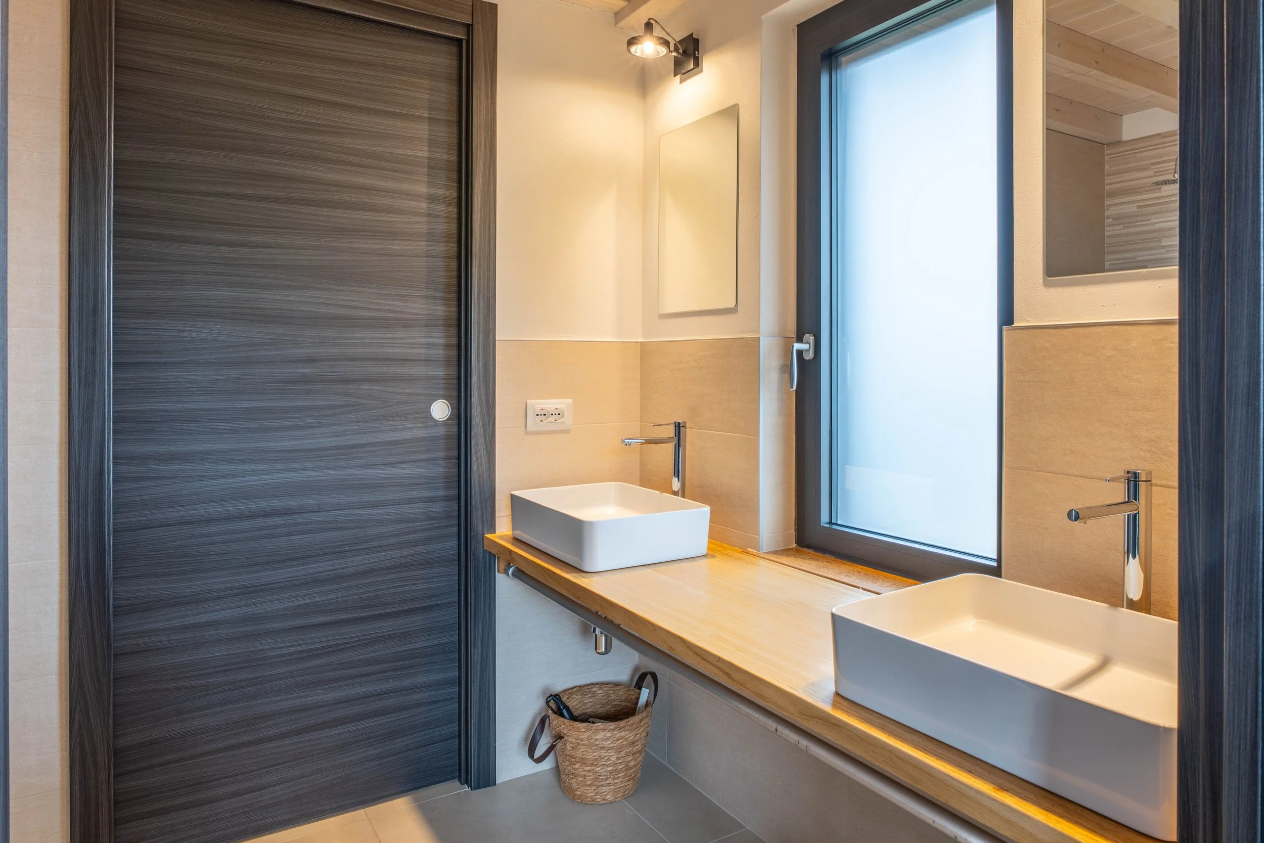 Moderne badkamer met twee vierkante wasbakken op een houten wastafel, een grote raam met frosted glas, een spiegel, een raam met een zwart frame en een muur met neutrale tinten.