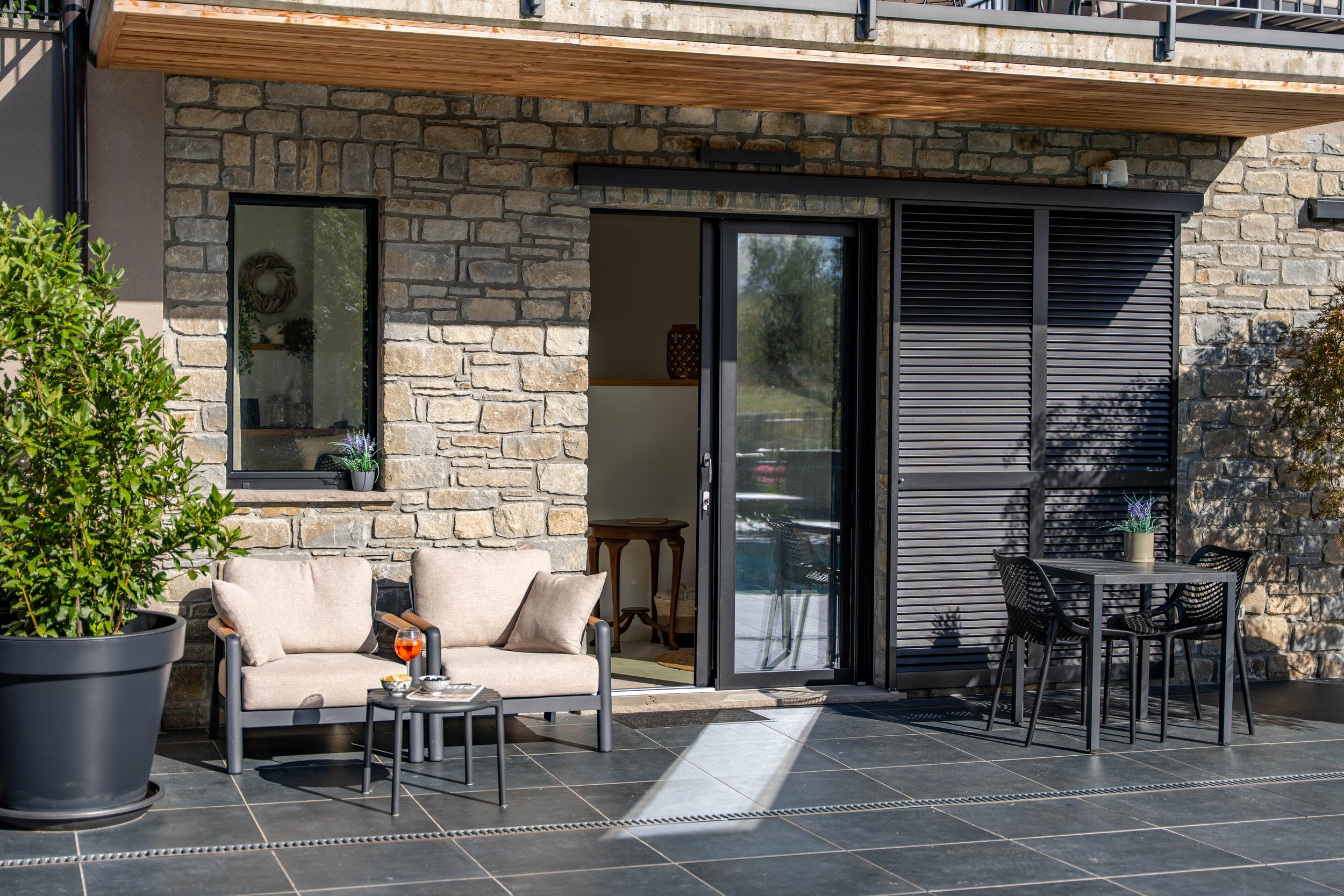 Een patio met een bankje en tafel, met een deur en ramen op een stenen muur, en een eettafel met stoelen buiten.