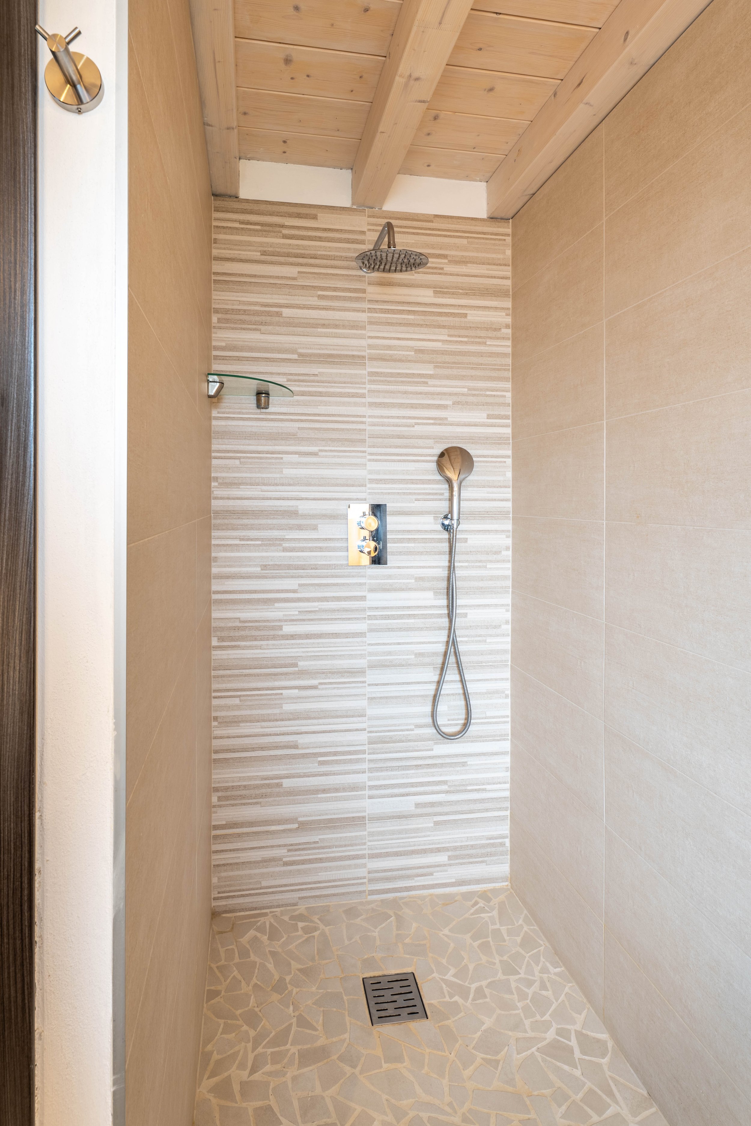 Een inloopdouche met beige en gestreepte wandtegels, een regen douchekop, een handdouche, een glazen plank en een afvoerputje op de vloer.