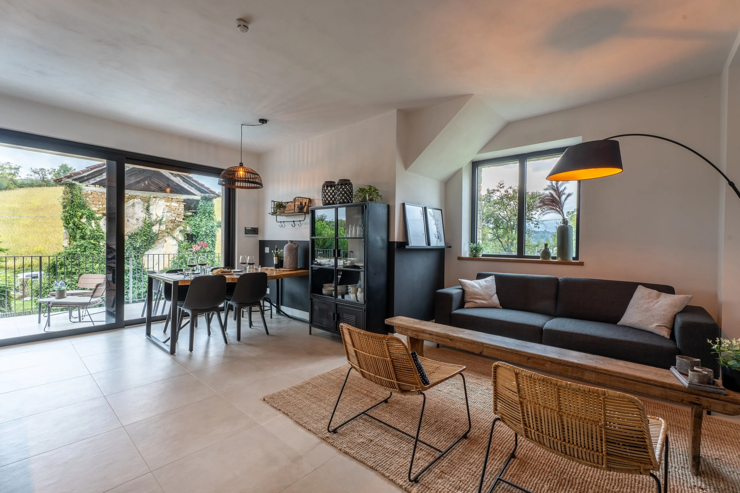Woonkamer en eetruimte met grote raampartijen, bank, stoelen, en een balkon met uitzicht op groen landschap.