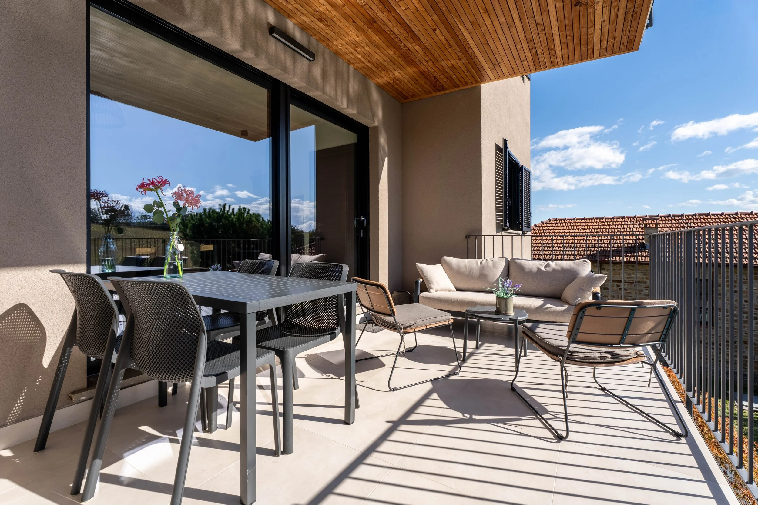 Balkon met buitentafel en stoelen, een bank en twee loungestoelen, met bloemen en uitzicht op een blauwe hemel met wolken.