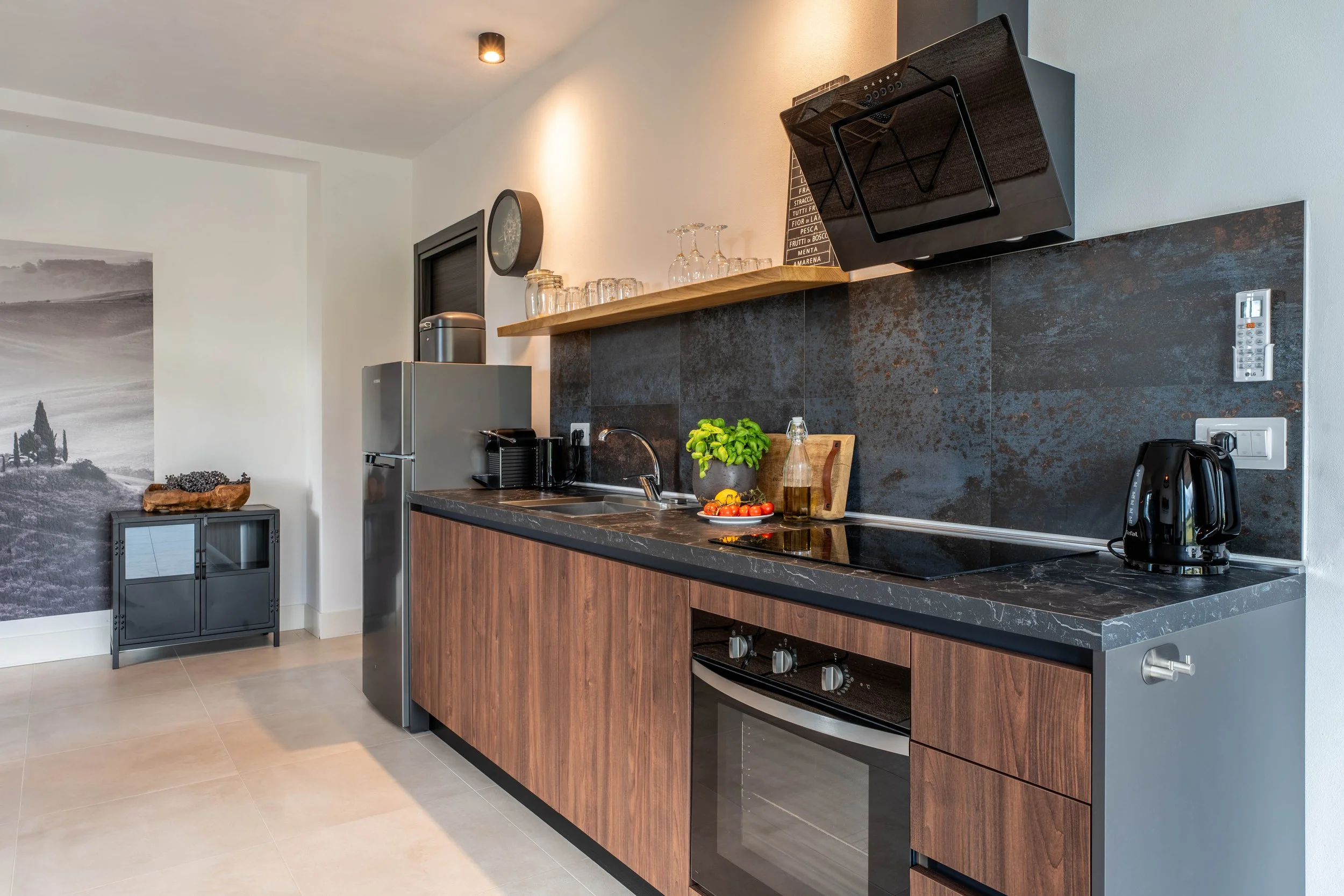 Moderne keuken met houten kasten en zwarte werkbladen, inclusief koelkast, oven, waterkoker, koffiezetapparaat, diverse glazen, een plantenpot, tomaten, citroen, fles olie en snijplank.