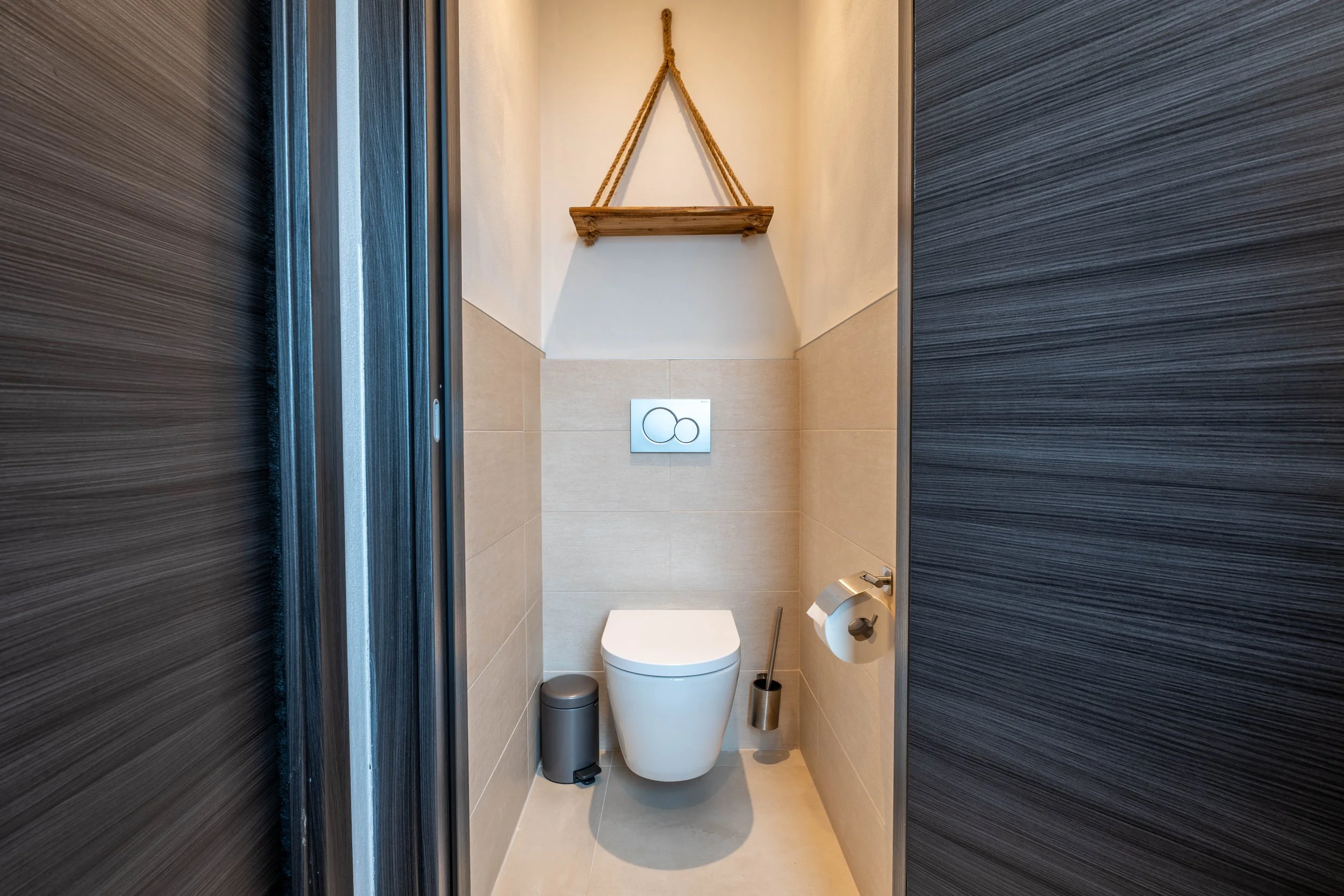 Toilet in een moderne badkamer met beige tegelwanden en donkere houten schuifdeuren, boven het toilet een houten plank met touwhangsysteem.