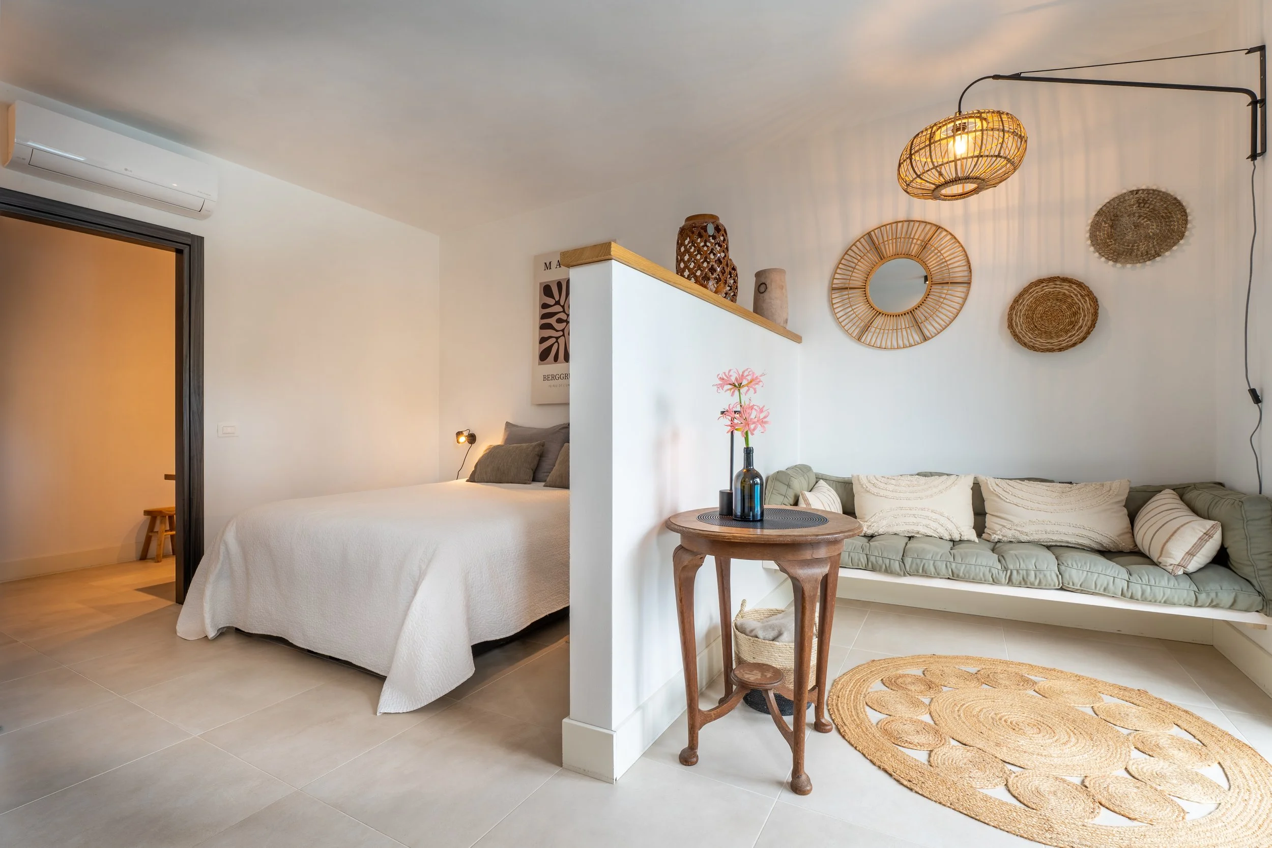 Interieur van een moderne slaapkamer met een tweepersoonsbed, een loungebank, decoratieve manden en spiegels op de muur, en een kleine houten tafel met bloemen.