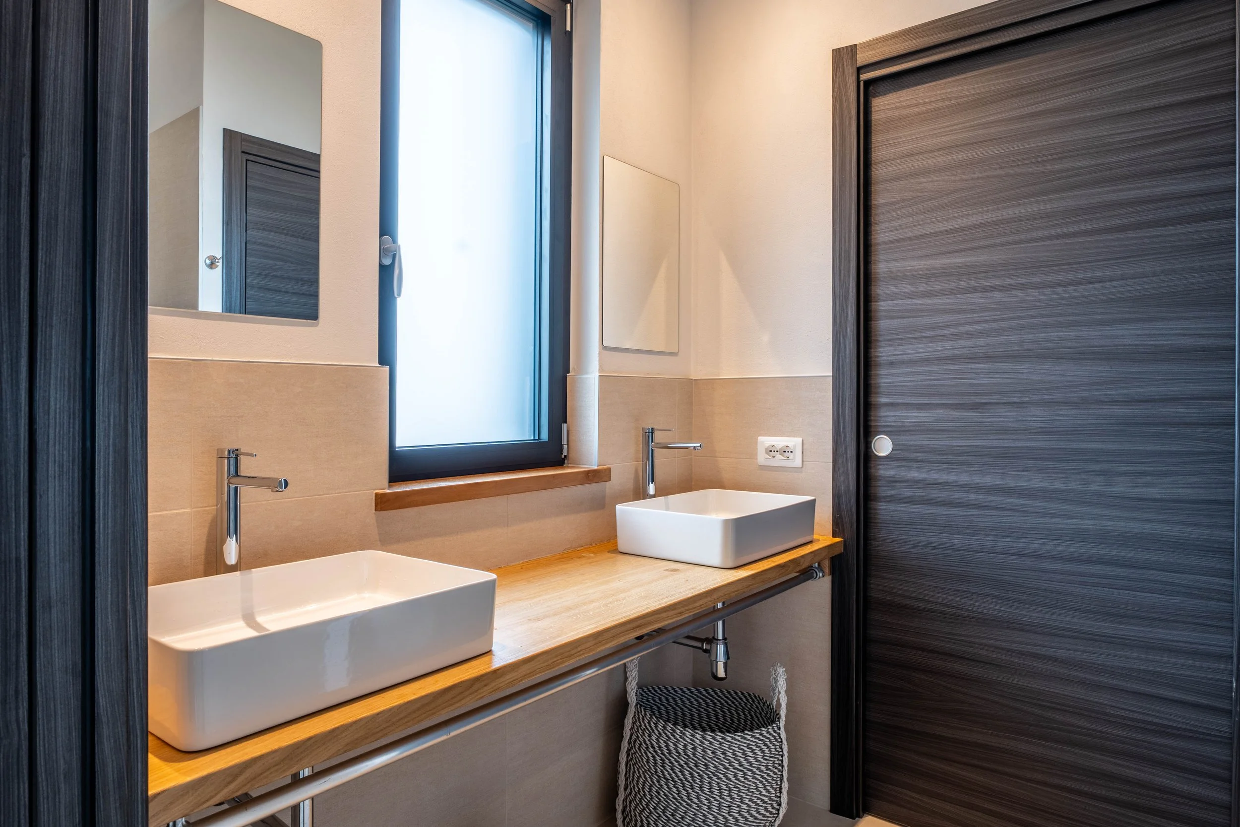 Tweede badkamer met twee wastafels, een raam met frosted glas, een spiegel, een wand met stopcontacten, en een opbergmand onder de wastafel.