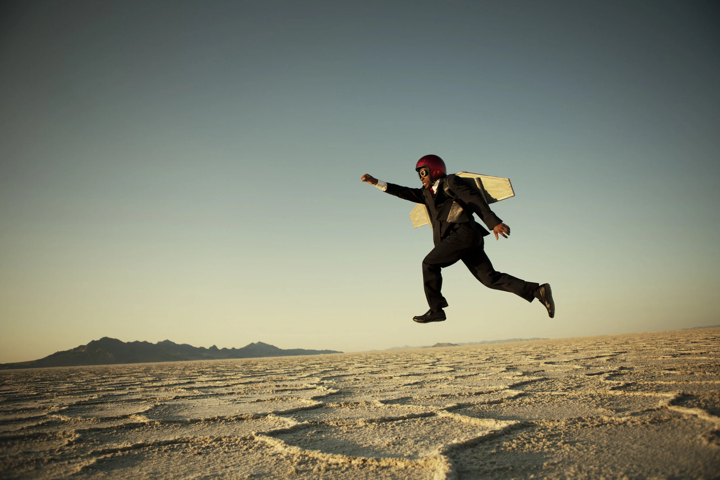 A man in a business suit, wearing a red helmet and goggles, is jumping in a desert landscape while carrying a cardboard airplane.