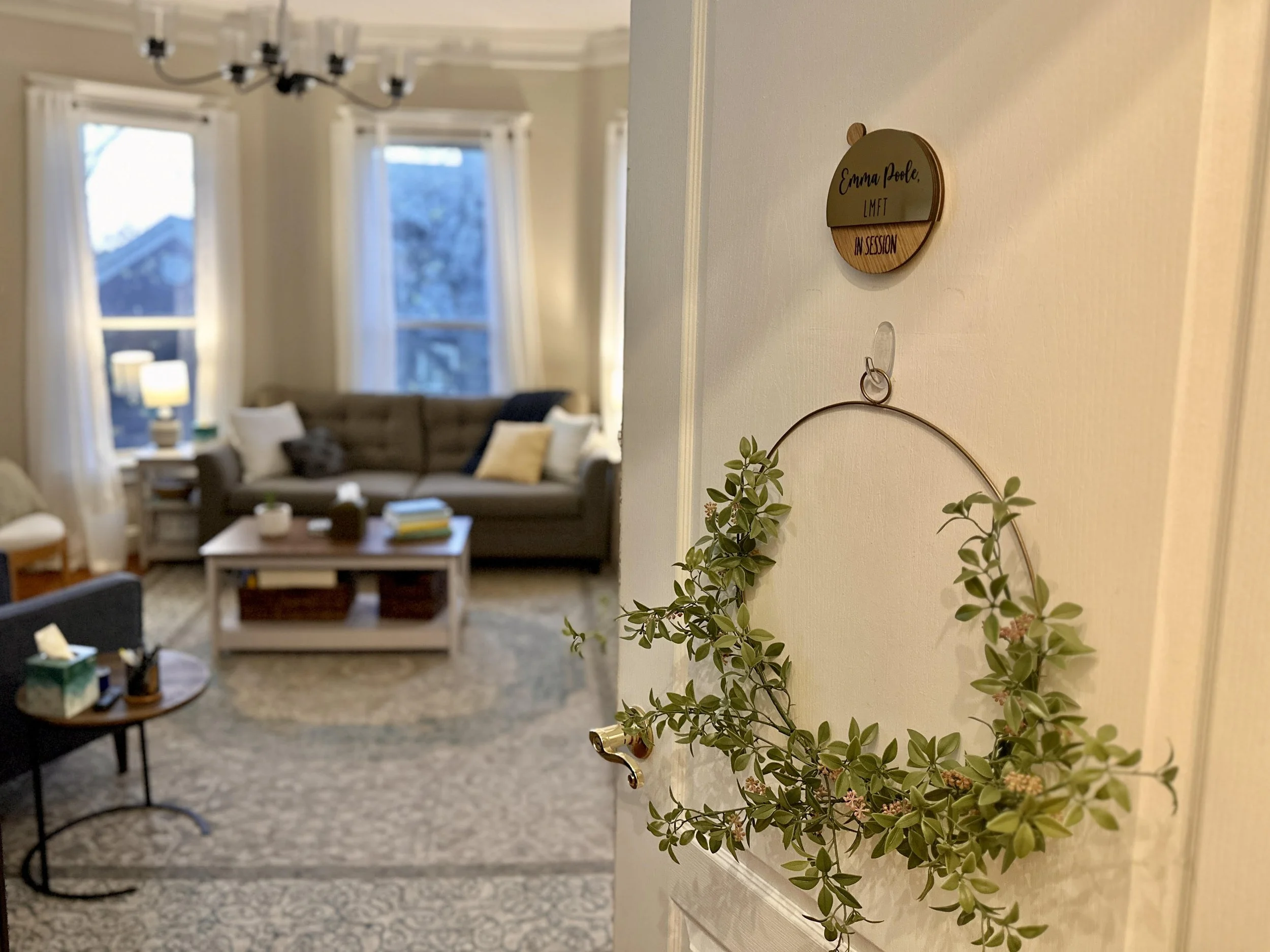 Entrance to therapy office with door decoration in focus. Our of focus background includes a cozy living room atmosphere with pillows, couches, books, coffee table, warm lighting.