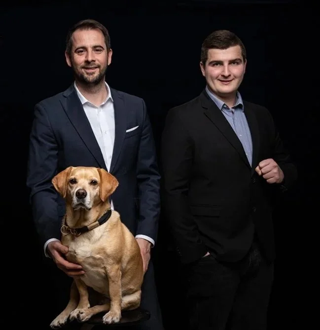 Zwei Männer in Anzügen stehen vor einem schwarzen Hintergrund, einer hält einen Hund in den Händen.