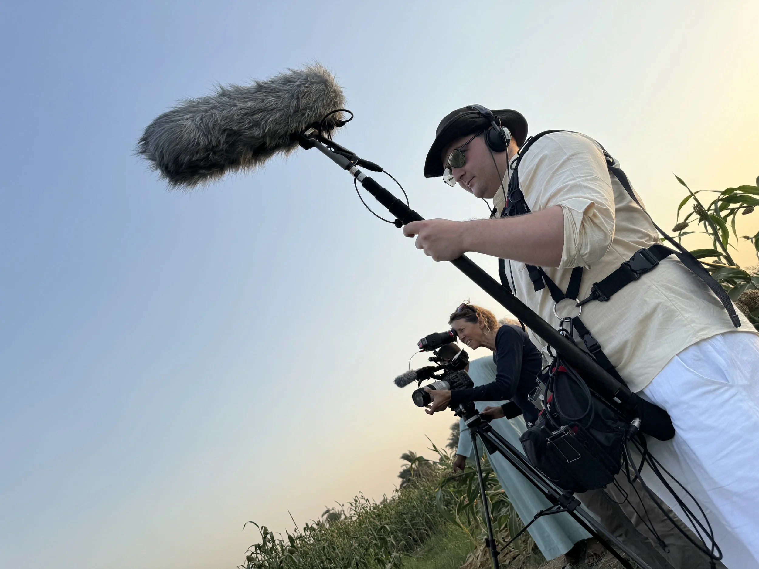 Zwei Menschen filmen in einer landwirtschaftlichen Gegend, einer mit einer Kamera und der andere mit einem Mikrofon, während sie den Himmel beobachten.