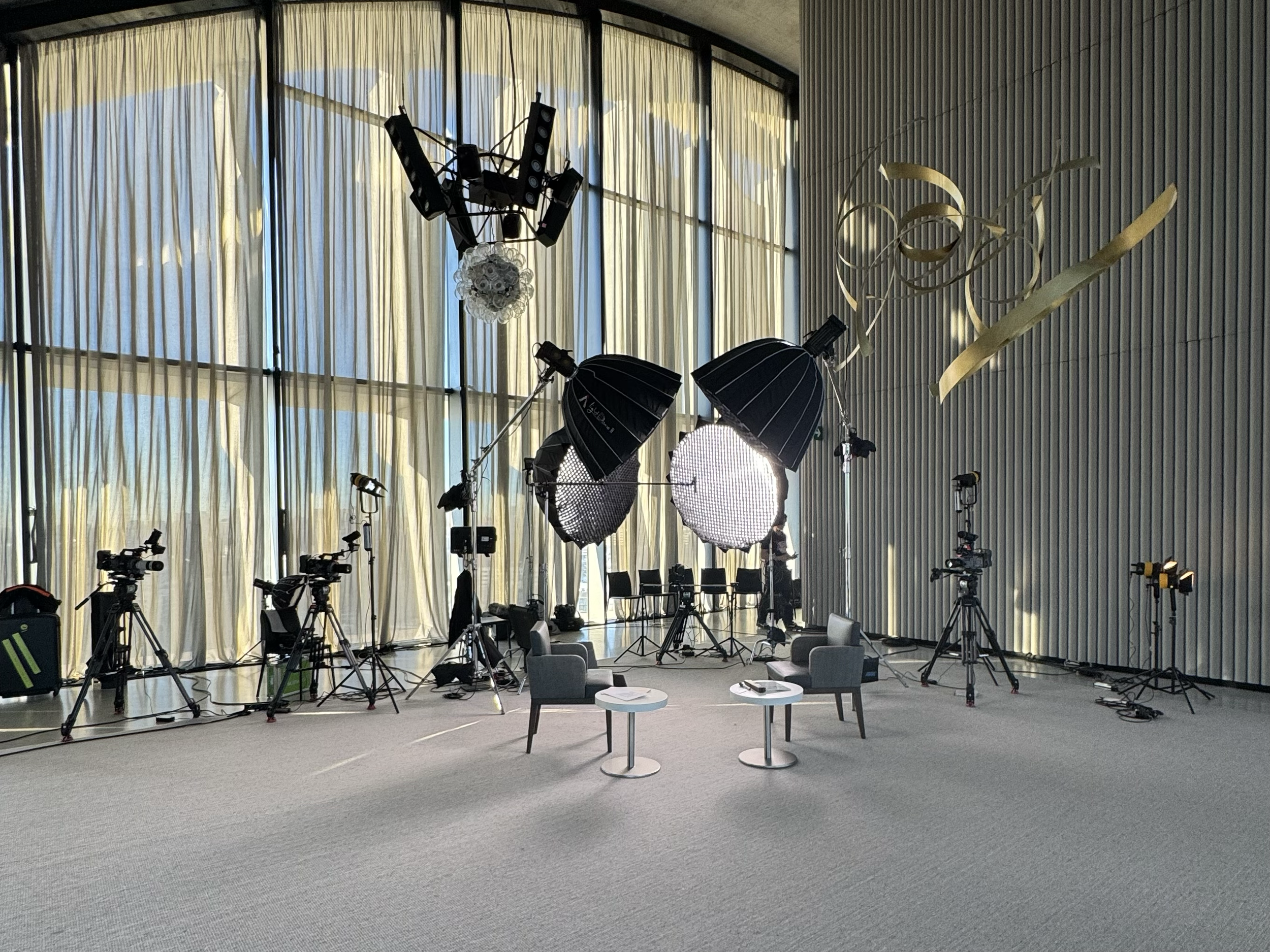 Fotostudio mit professioneller Beleuchtungs- und Kameratechnik, zwei Stühle und kleine Tische, große Fensterfront mit Vorhängen.