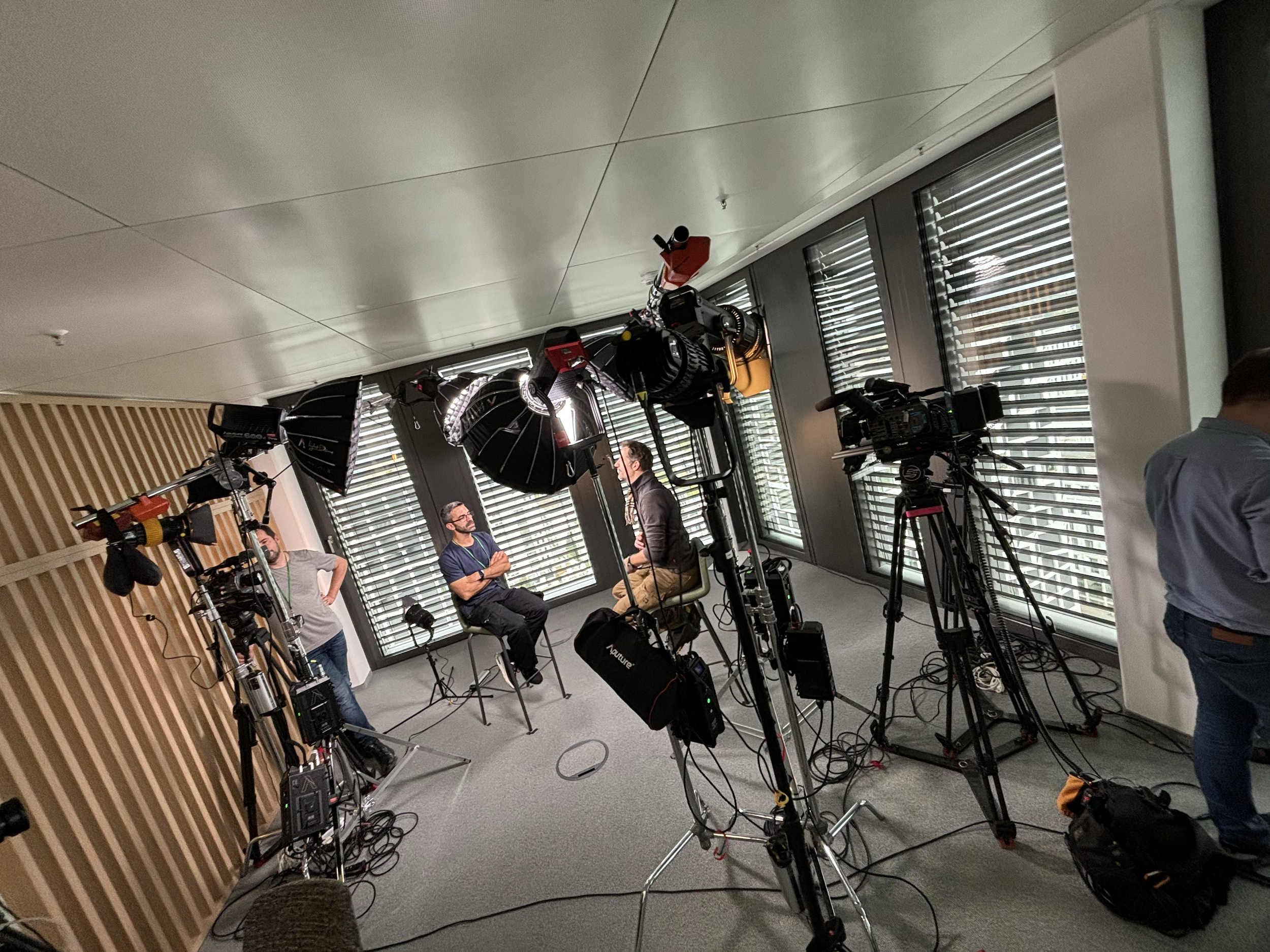 Interview im Studio mit zwei Männern, Kamera, Beleuchtung, und technische Ausrüstung, während des Gesprächs an den Fenstern mit Rollläden.