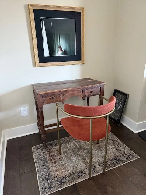 A framed photograph of Chris Farley by Chris Buck hangs in a wall above a small wood desk. A red and gold chair sits empty beside the desk facing the photograph.