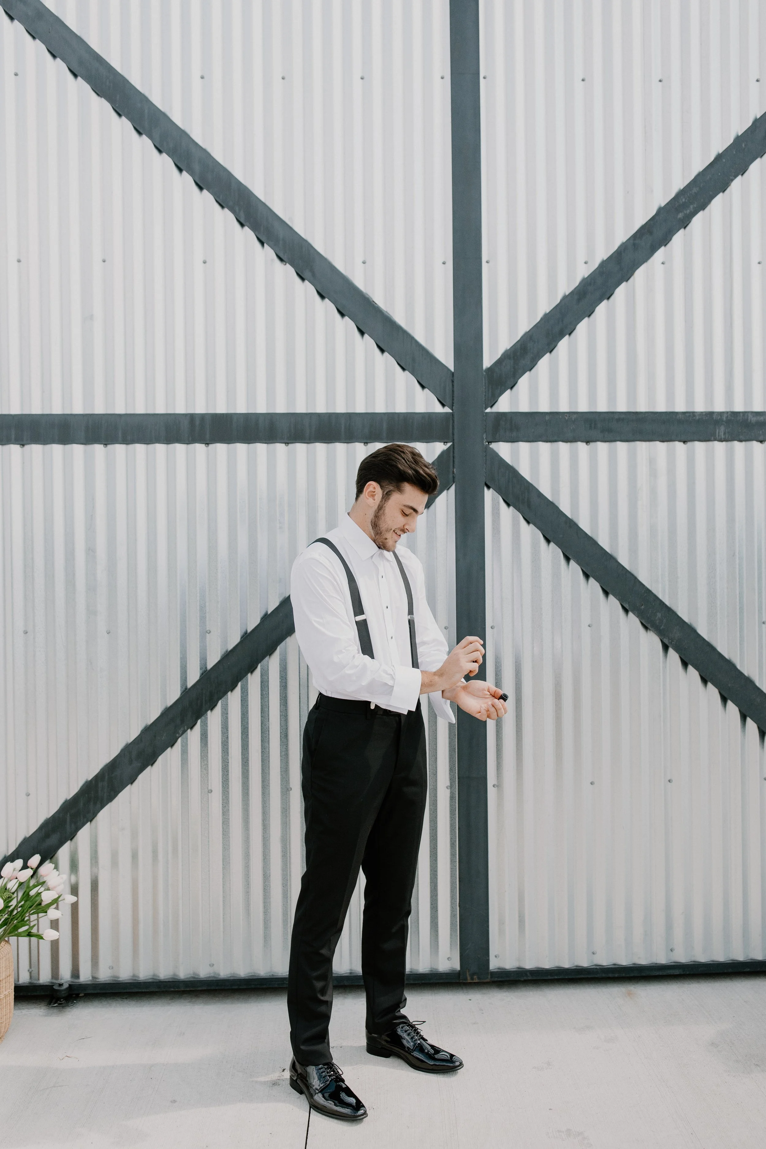A man dressed in a white shirt and black pants with suspenders, standing against a metallic industrial wall, holding and looking at a small object in his hands. This man is getting ready to get married.