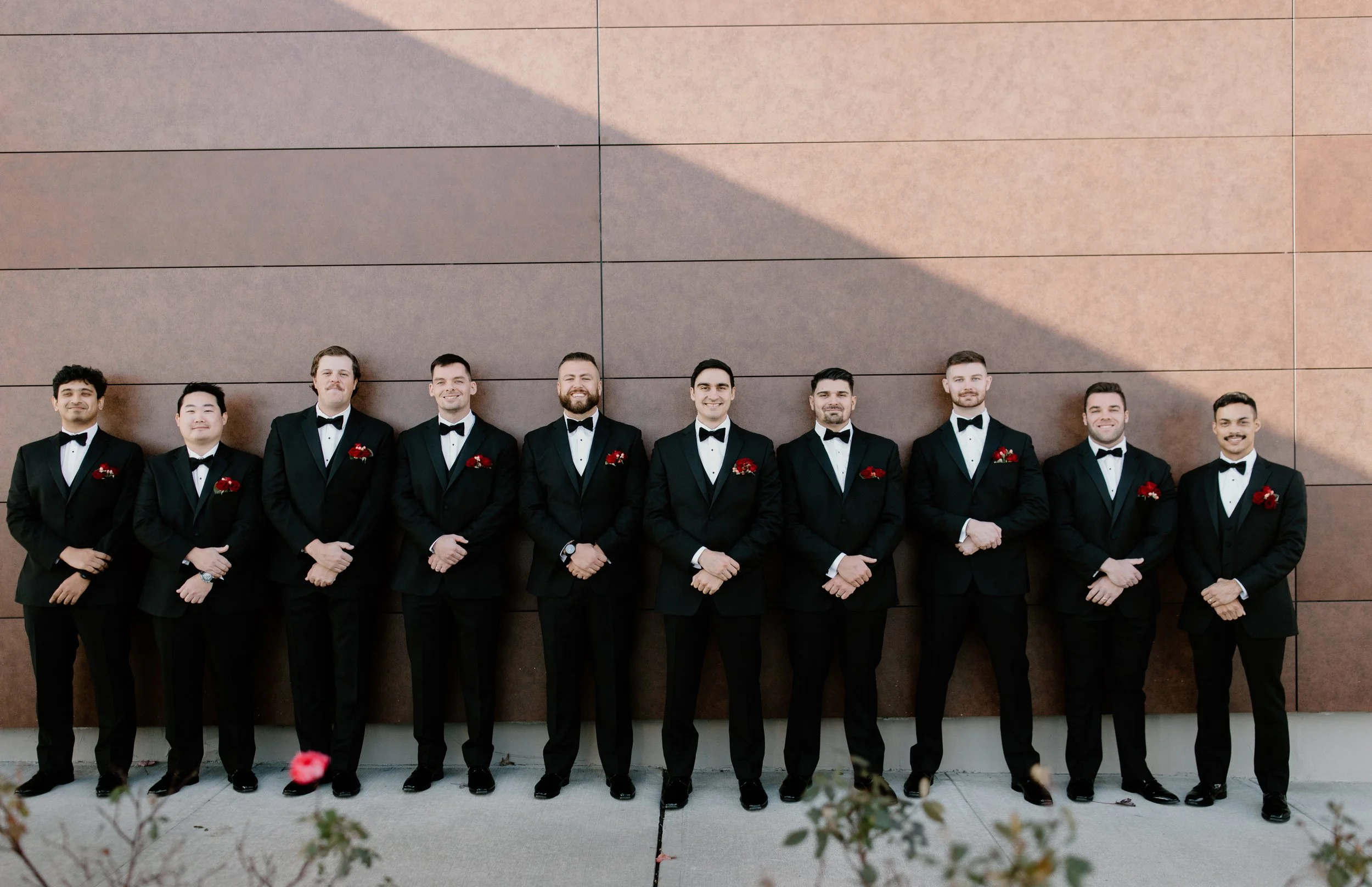 Group of eleven men in black tuxedos with bow ties, standing against a brown tiled wall, dressed for a formal event.