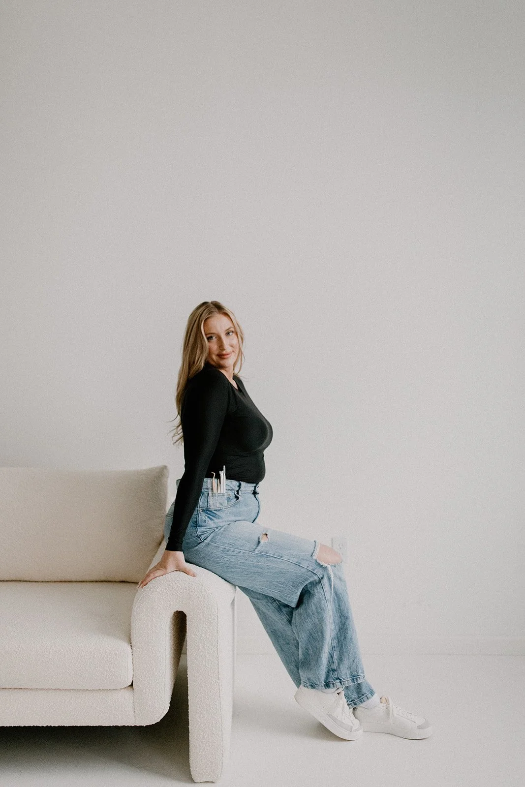 A woman in a black long-sleeve shirt and ripped jeans sitting on the armrest of a cream-colored sofa in This Hudson Studio in Wappingers NY  with a plain white wall.