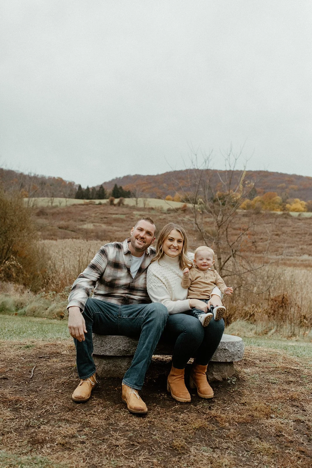 A family of three sitting on a stone bench outdoors in Danbury CT with a fall landscape, with mountains and trees with autumn foliage in the background.