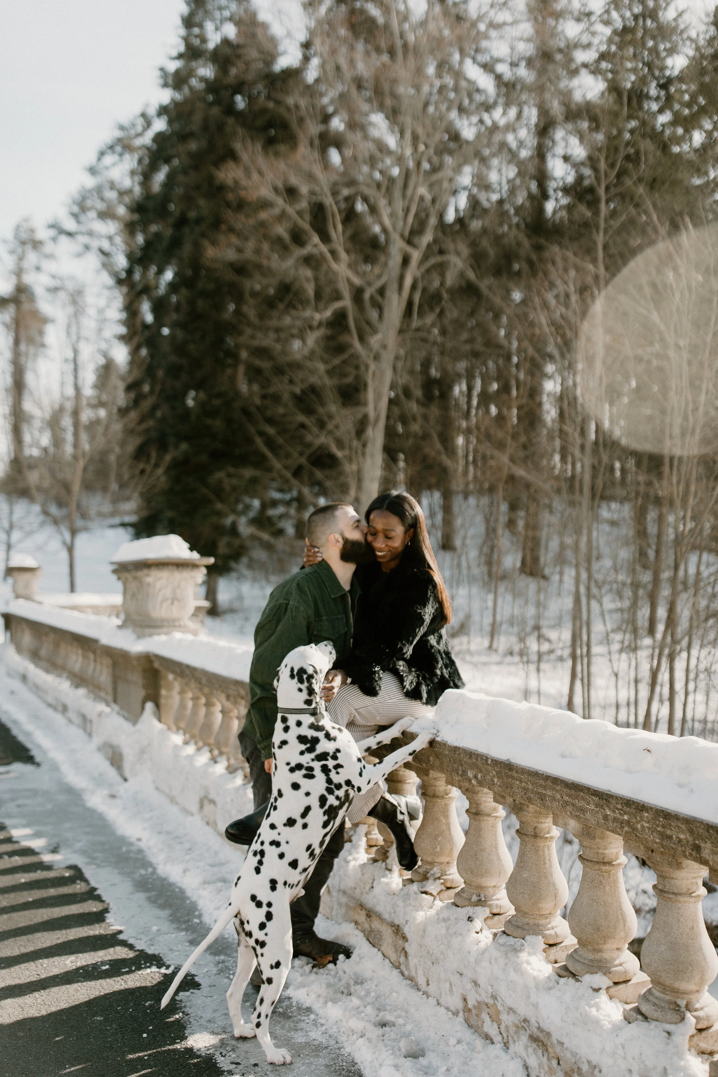A couple and a Dalmatian dog enjoying a snowy day outdoors, with the woman sitting on a stone railing and the man standing nearby.
