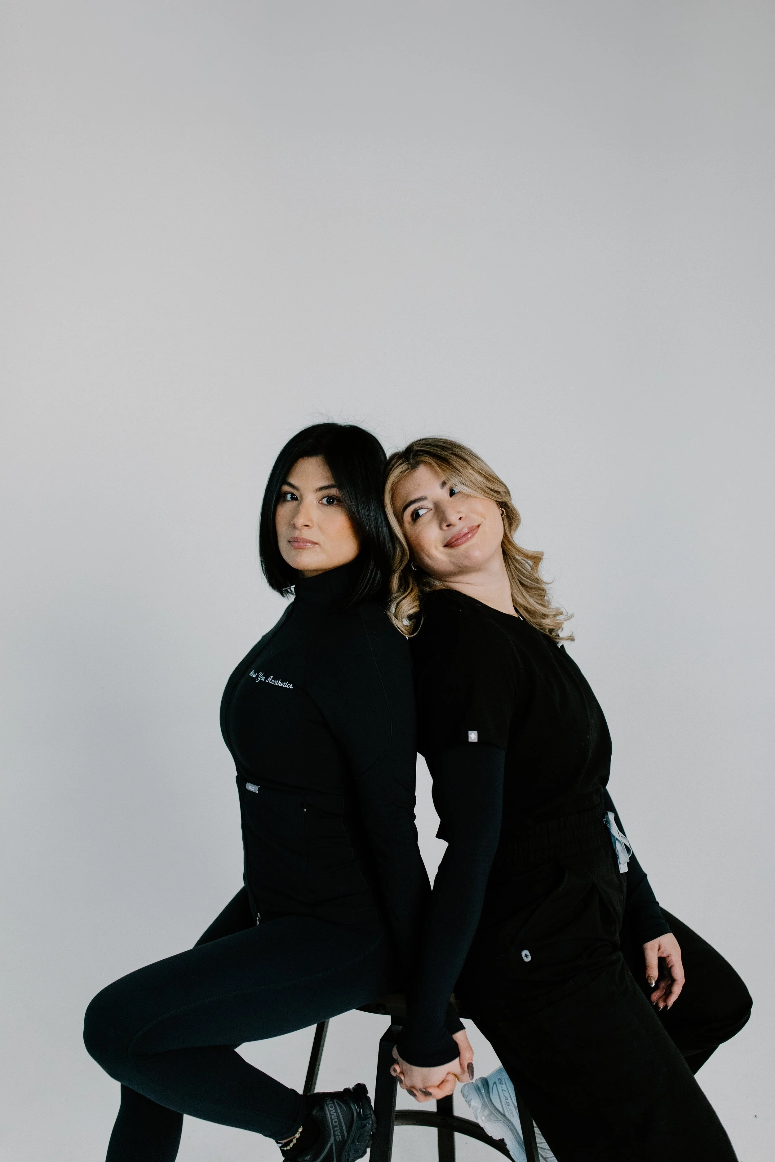 Two women sitting back to back on a stool, holding hands, against a plain white background. The woman on the left has black hair and an athletic outfit, while the woman on the right has blonde hair and a black outfit.