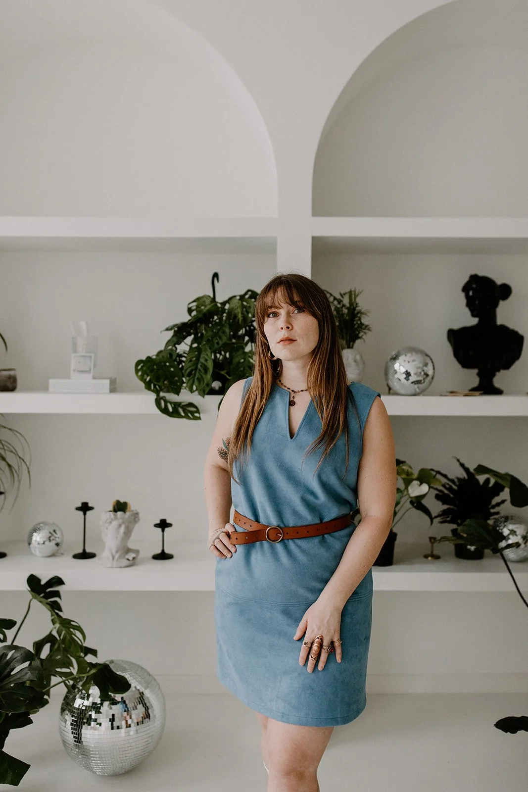 A woman with long brown hair and bangs is standing inside This Hudson Studio. She is standing in front of white wall shelves decorated with plants and art objects. She wears a sleeveless blue dress with a brown belt and has rings and a necklace.