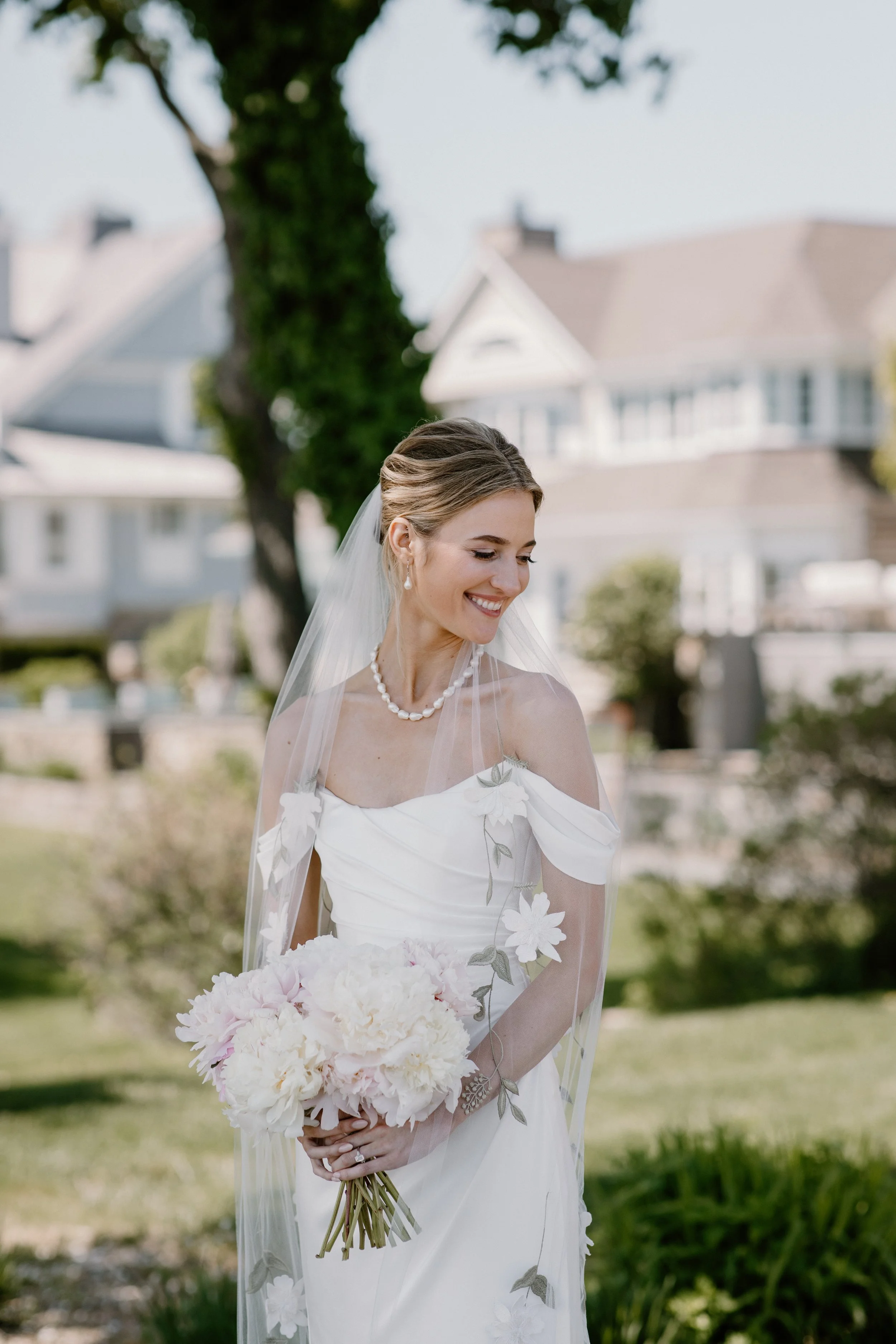 A bride in a white wedding dress holding a bouquet of flowers outdoors with houses and trees in the background. A beach wedding in Westport CT