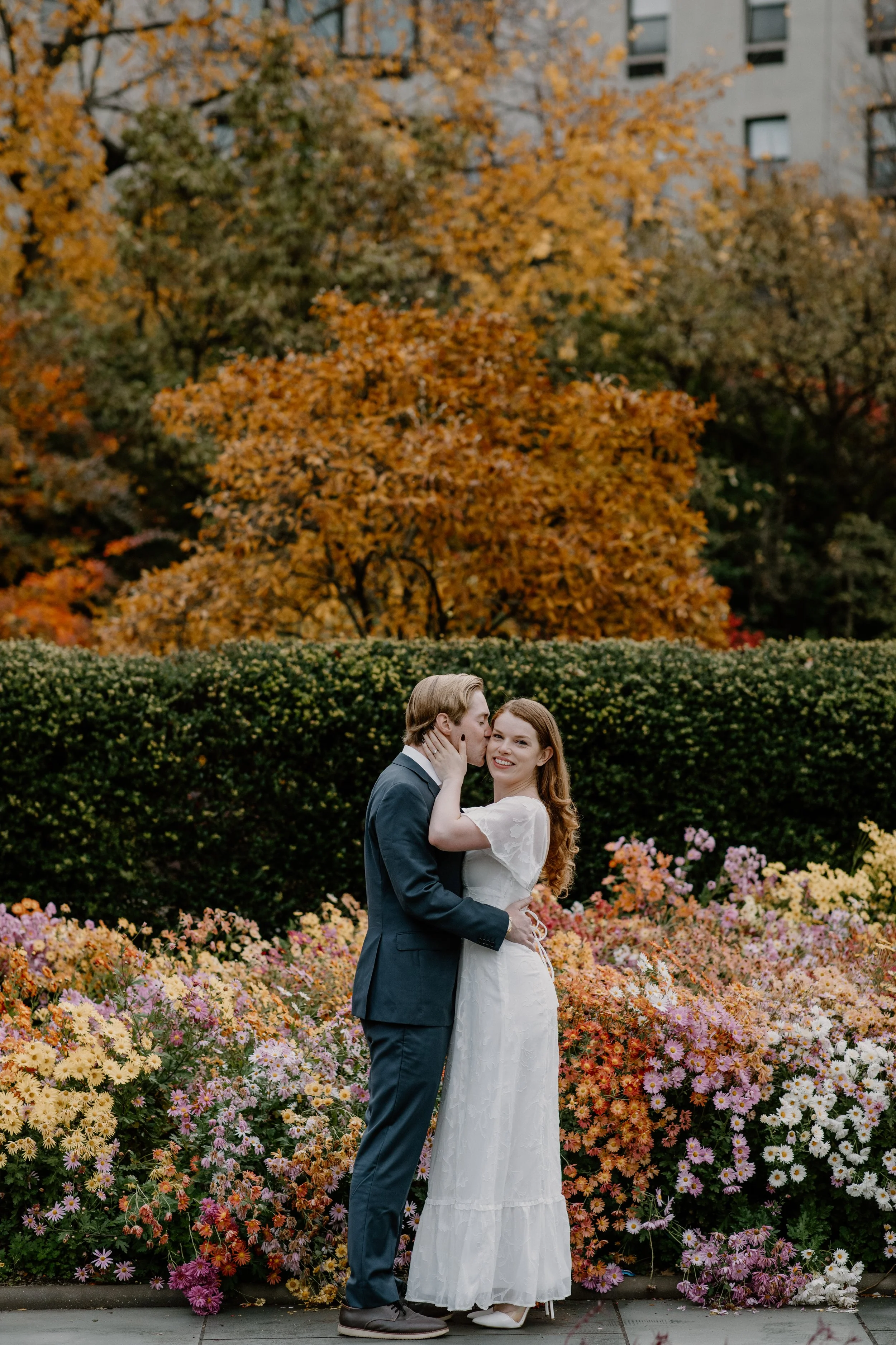 A couple wearing wedding attire standing in a garden with colorful flowers in Central Park. Autumn trees in the background, sharing a kiss and smiling.