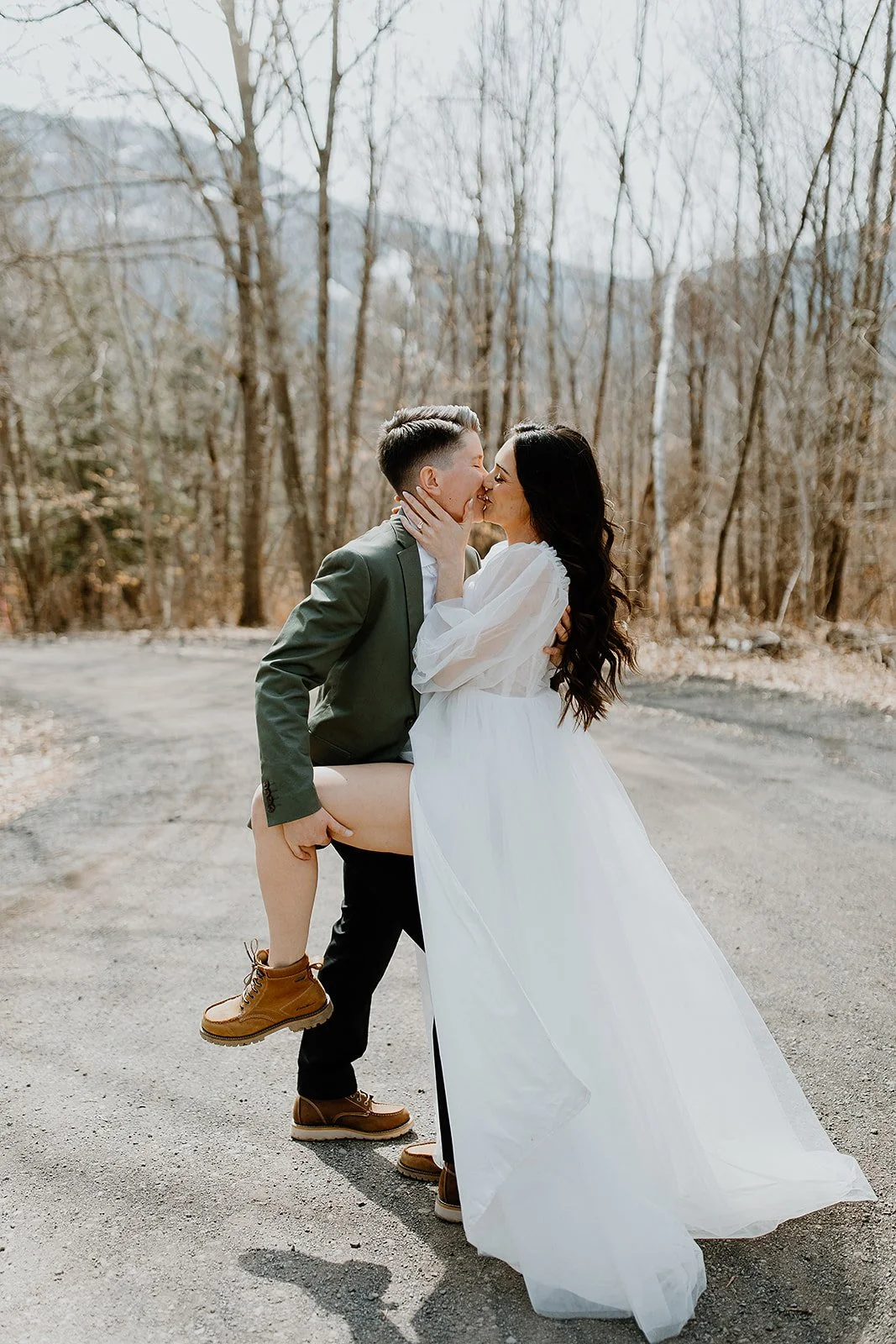 A couple dressed in wedding attire sharing a kiss outdoors on a dirt road, with leafless trees in the background.