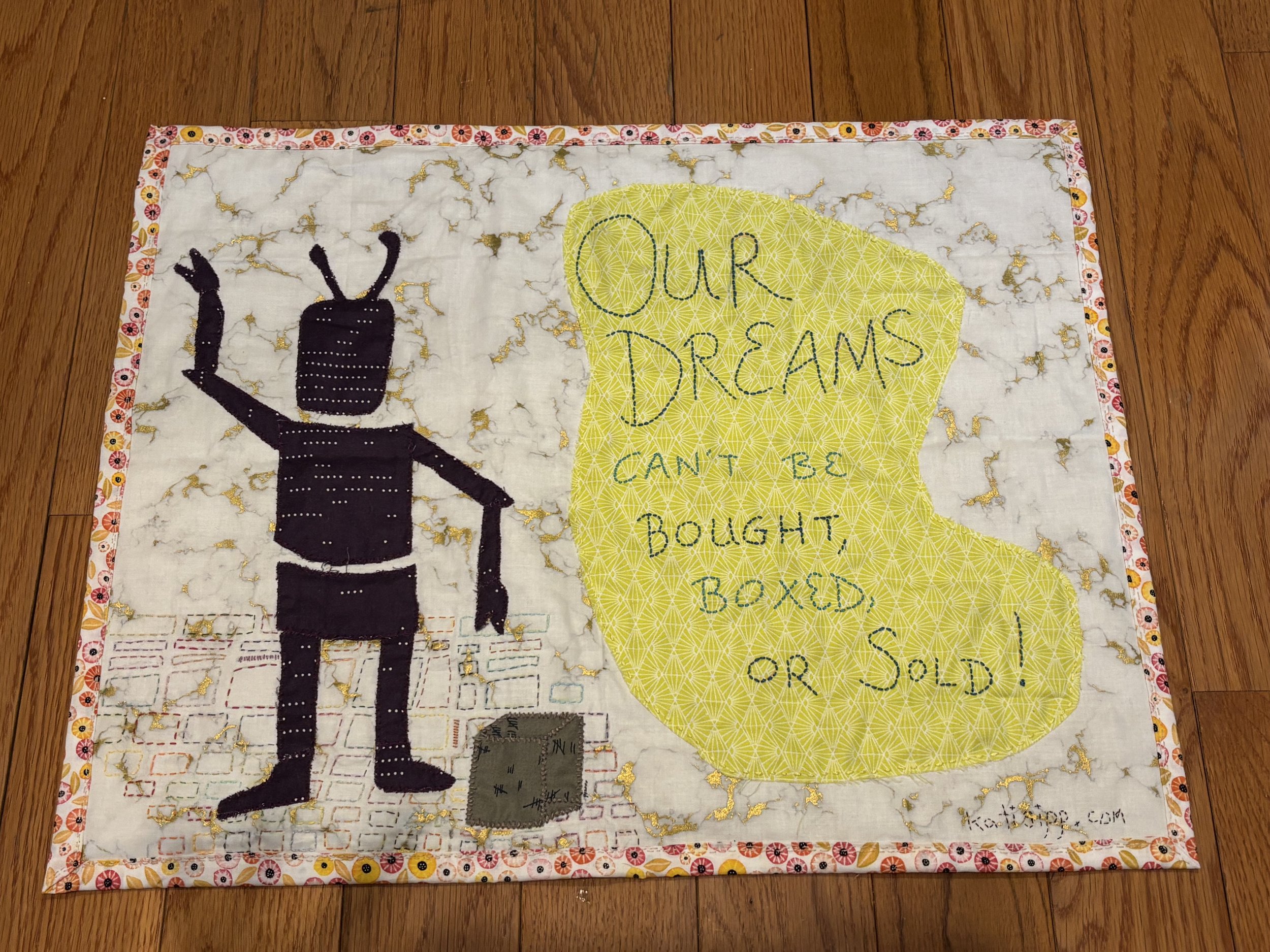 a fabric wall hanging with an image of a robot, some boxes, and "Our Dreams Can't Be Bought, Boxed, or Sold" embroidered