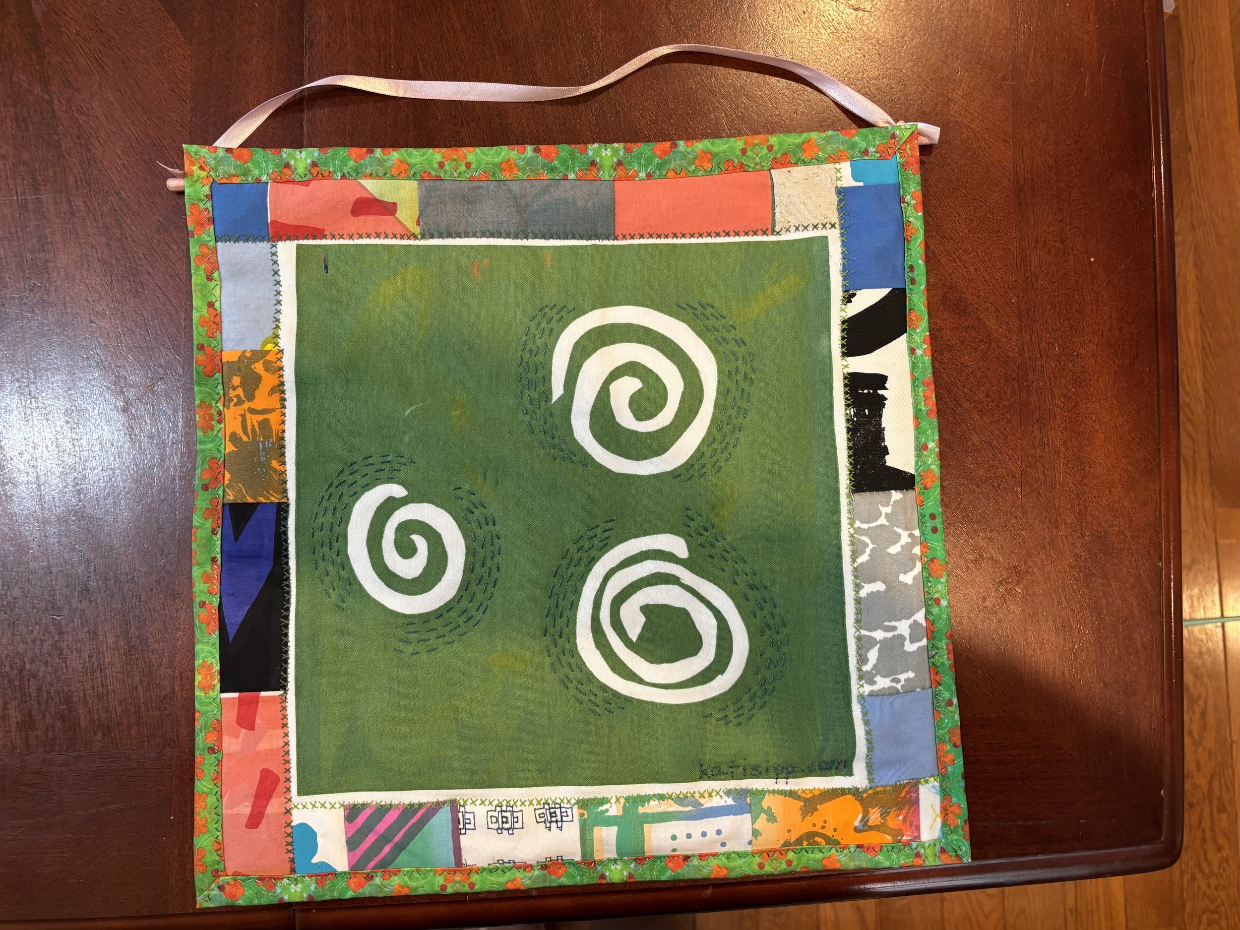 a fabric wall hanging that shows three hand-drawn spirals. The spirals are white on a green background, with a multi-colored border.