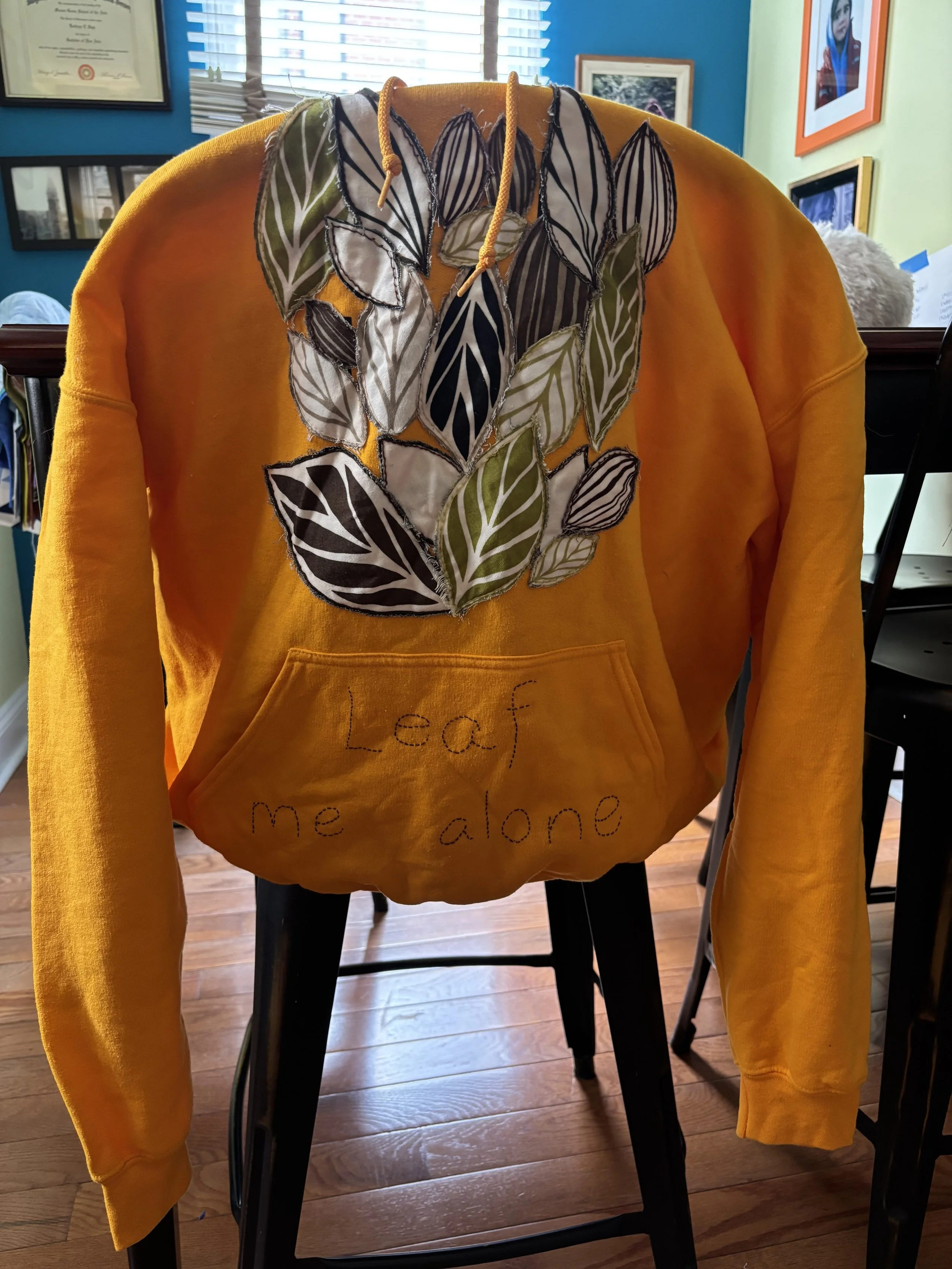 image of a yellow hooded sweatshirt with green and brown leaves appliquéd on it, and the words "Leaf Me Alone" embroidered on the pocket