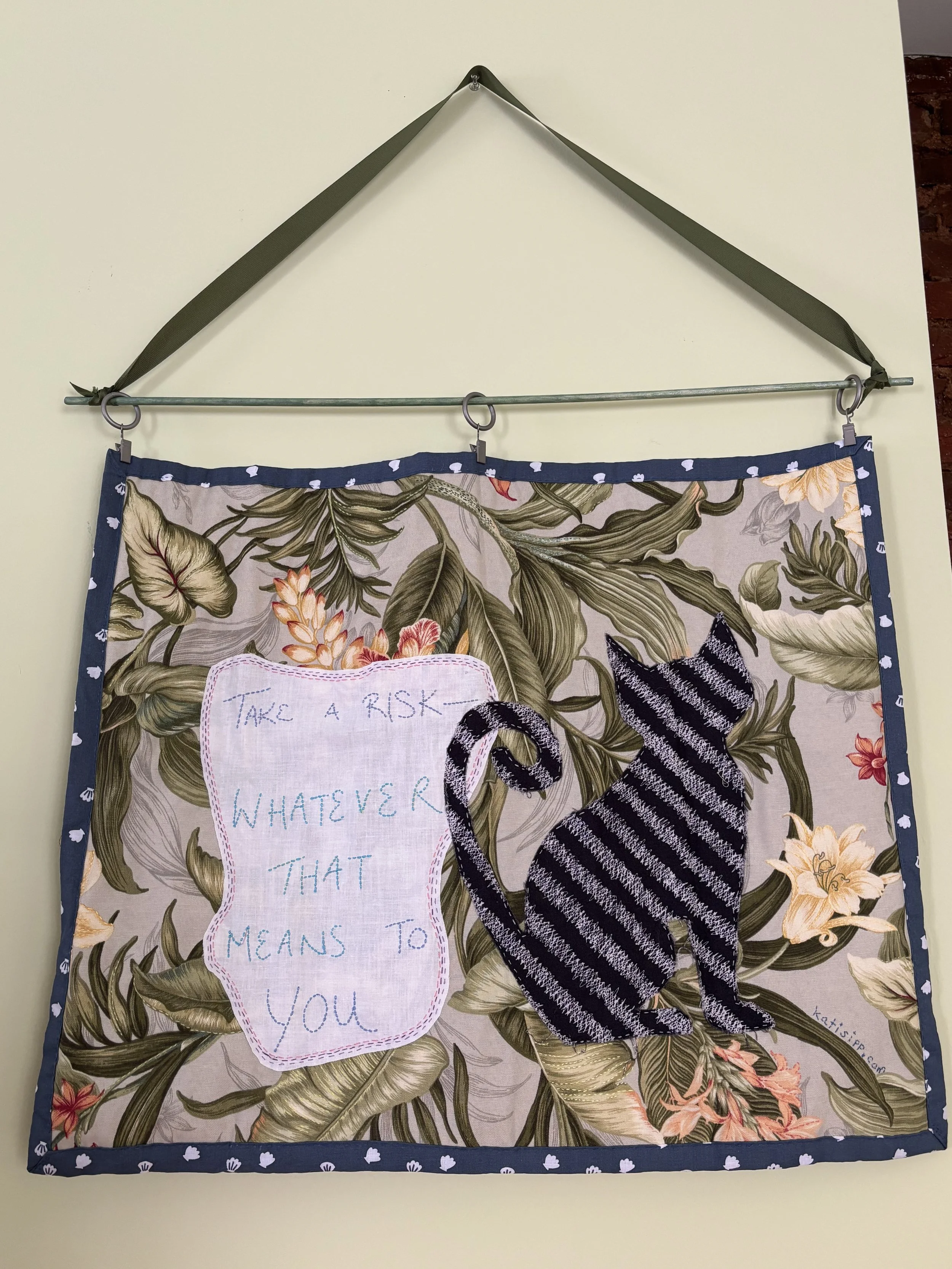 a fabric wall hanging that depicts a grey-striped black cat, and is embroidered with "take a risk, whatever that means to you"