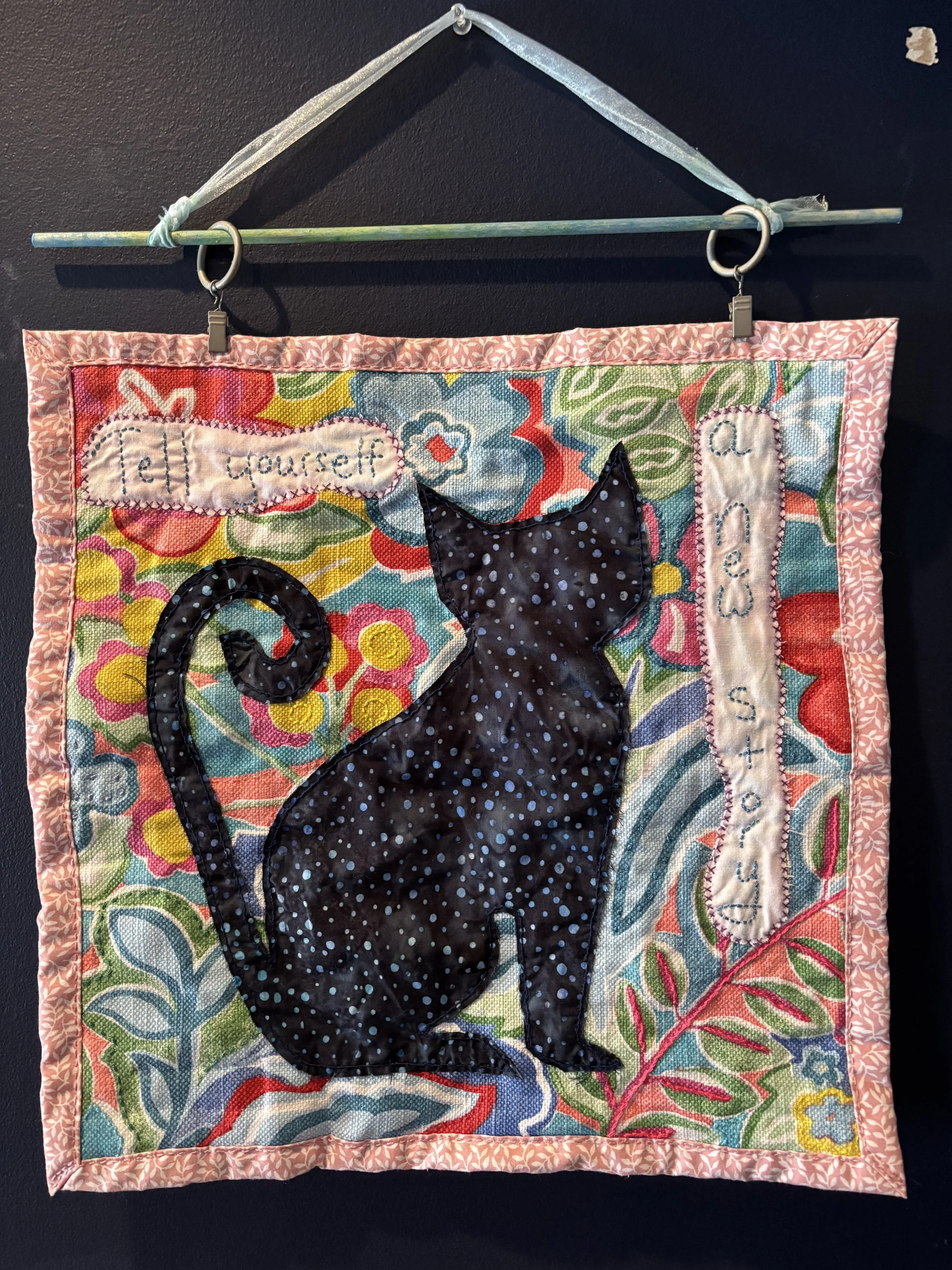 a fabric wall hanging that reads "Tell yourself a new story" and depicts a black cat silhouette on a background of colorful flowers