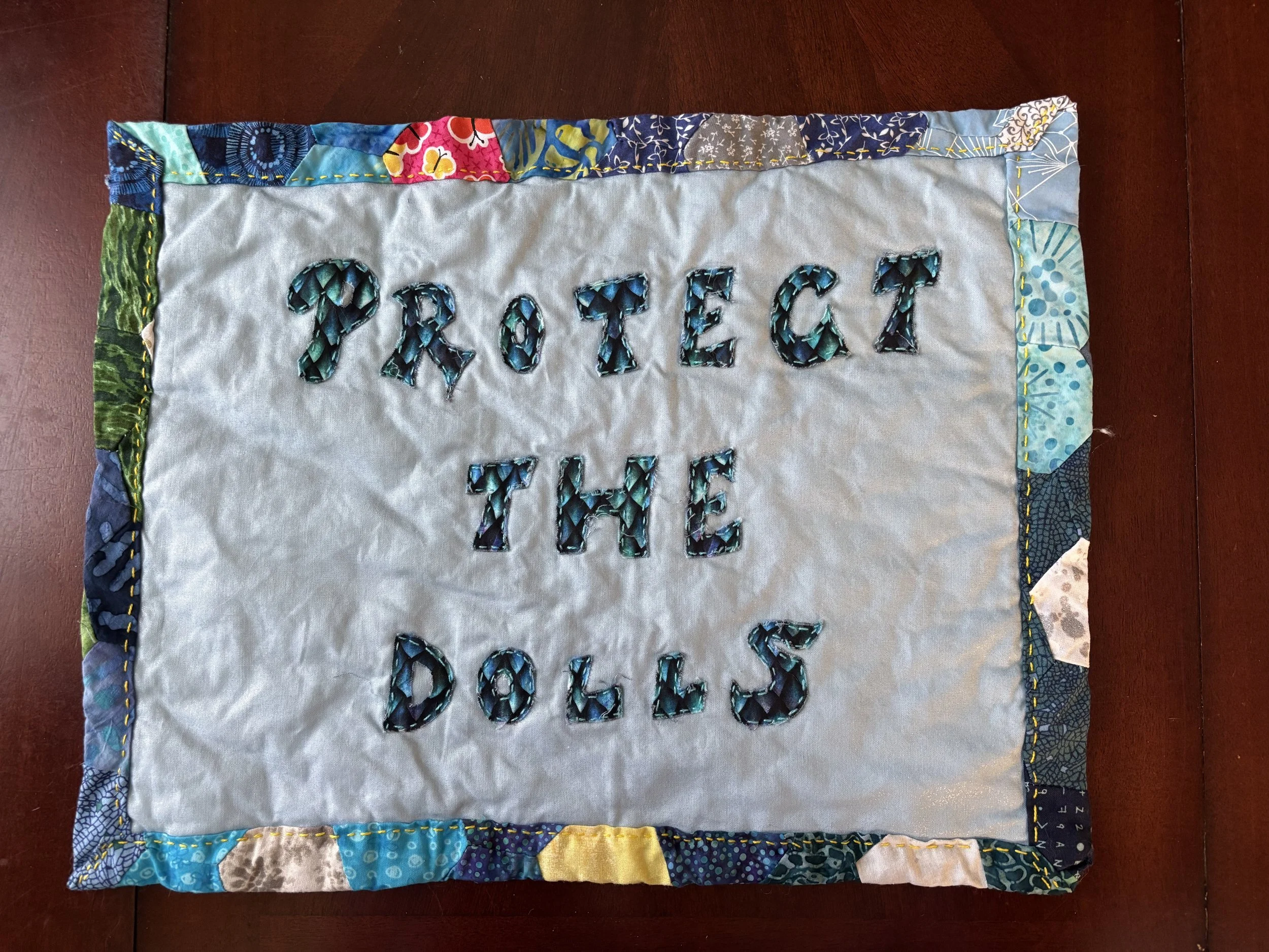 fabric wall hanging with "Protect the Dolls" appliquéd on the front
