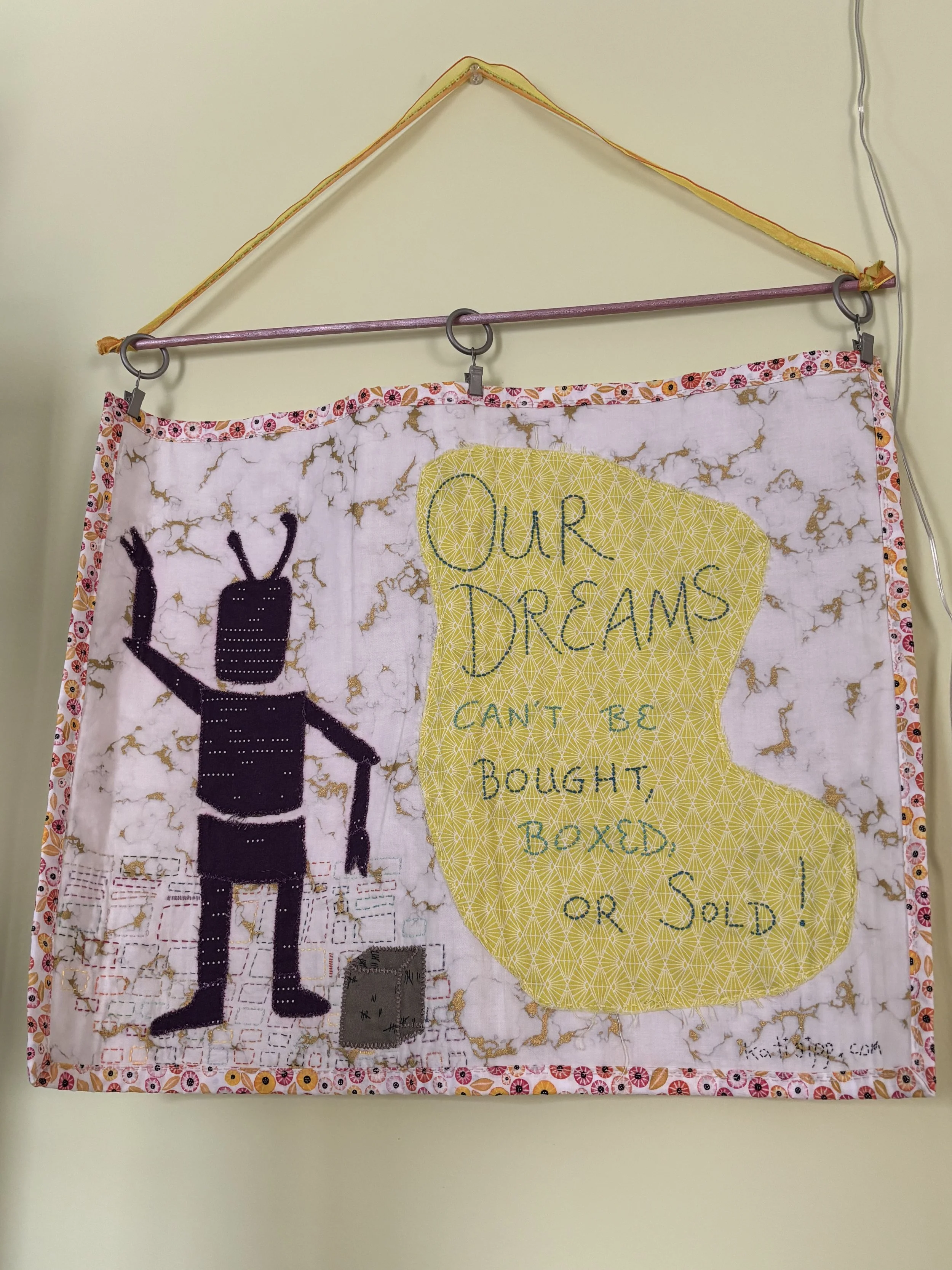 a fabric wall hanging with an image of a robot, some boxes, and "Our Dreams Can't Be Bought, Boxed, or Sold" embroidered