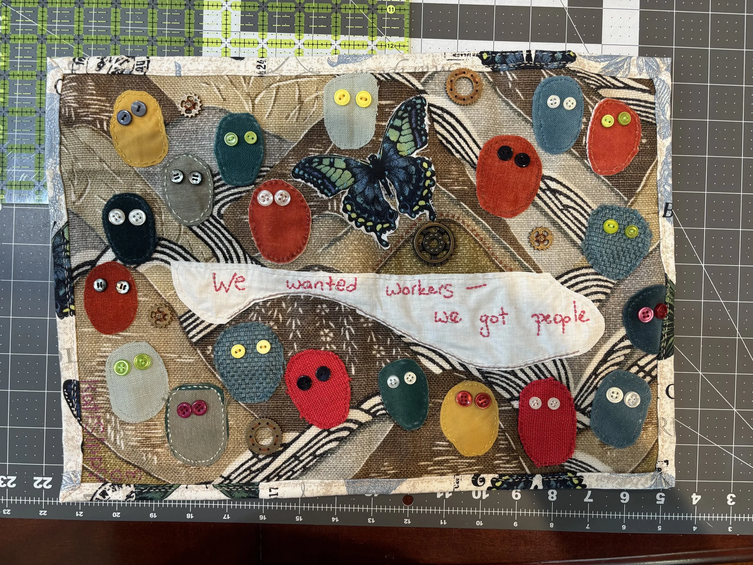 a hand-sewn fabric wall hanging that reads "We wanted workers -- we got people. There are many colorful "people" faces with eyes made of buttons.