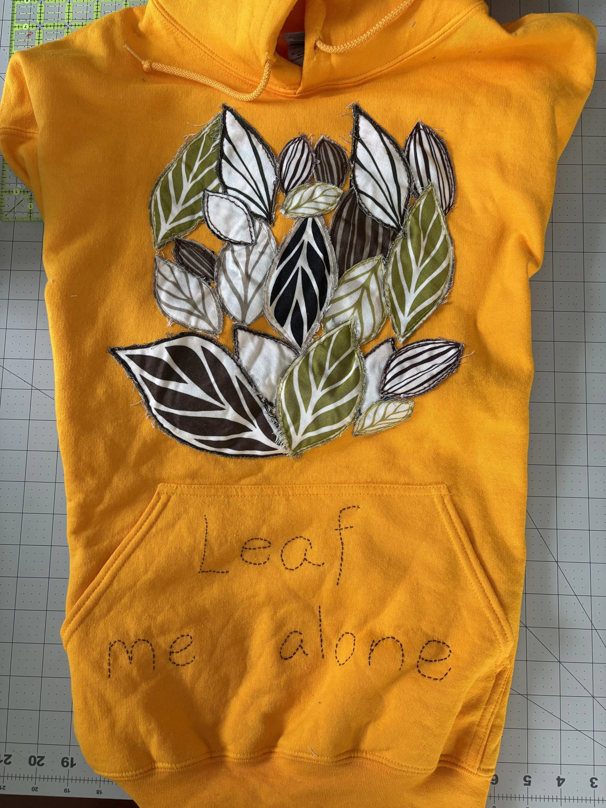 an upcycled yellow hoodie with green & brown leaves appliquéd on it, and "Leaf me alone" on the pocket