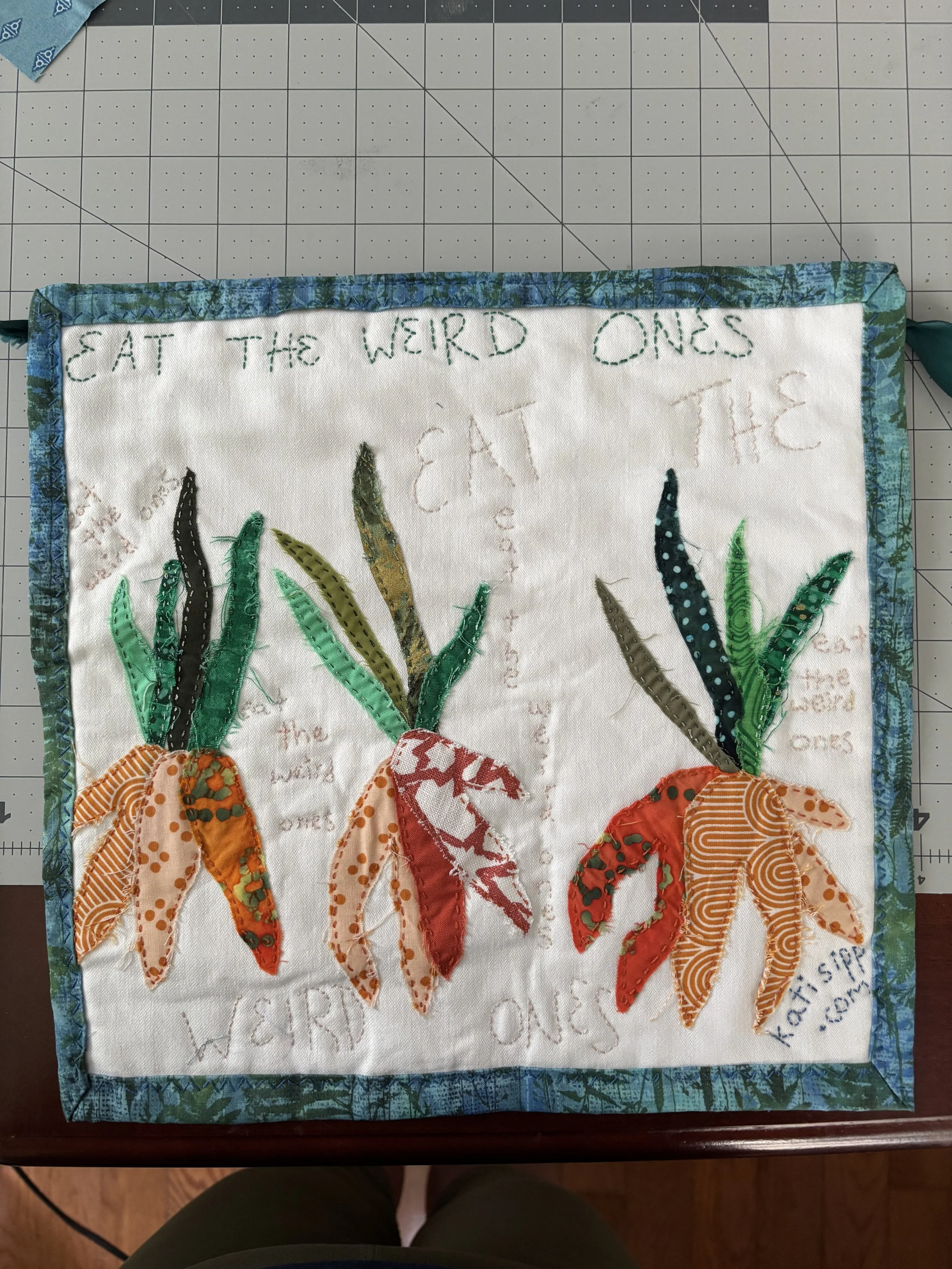 a fabric wall hanging that shows three bunches of odd carrots, and reads "eat the weird ones"