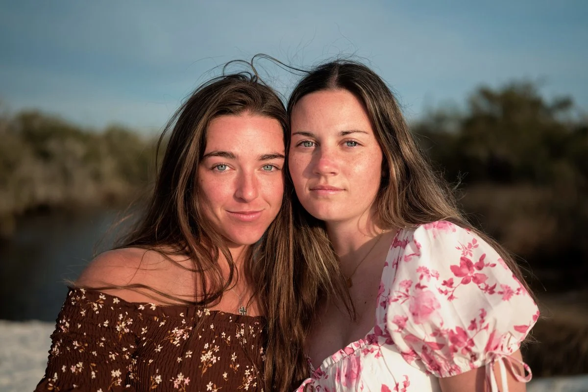 Two young women with long hair and light skin, one wearing a floral brown off-shoulder top and the other in a white floral dress, posing closely outside during daytime with a blurred outdoor natural background.